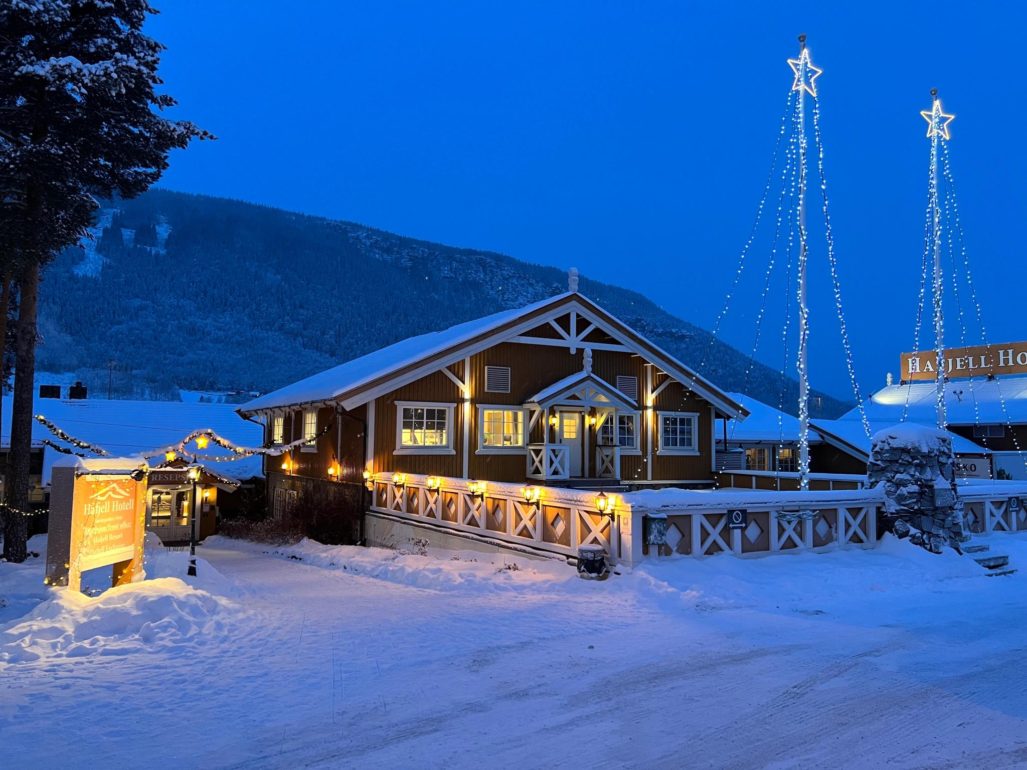 Upplyst träbyggnad i snö med stjärnformade ljus, berg i bakgrunden och blå skymningshimmel.