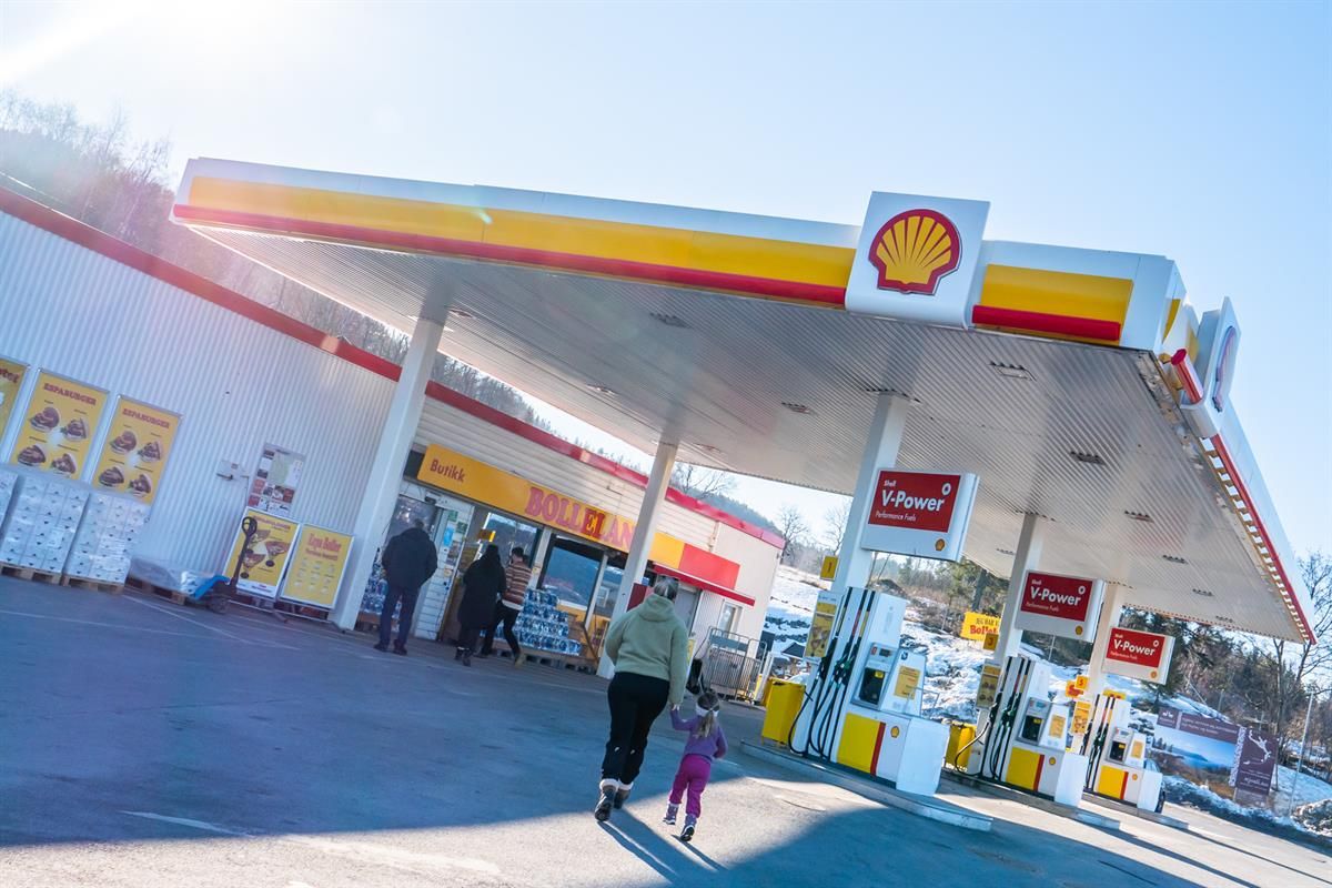 A Shell-branded gas station where customers walk between the fuel pumps and the store entrance on a sunny day.