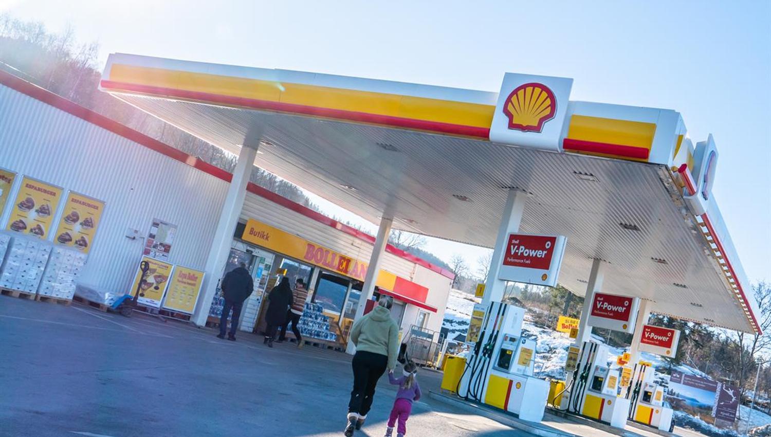 A Shell-branded gas station where customers walk between the fuel pumps and the store entrance on a sunny day.