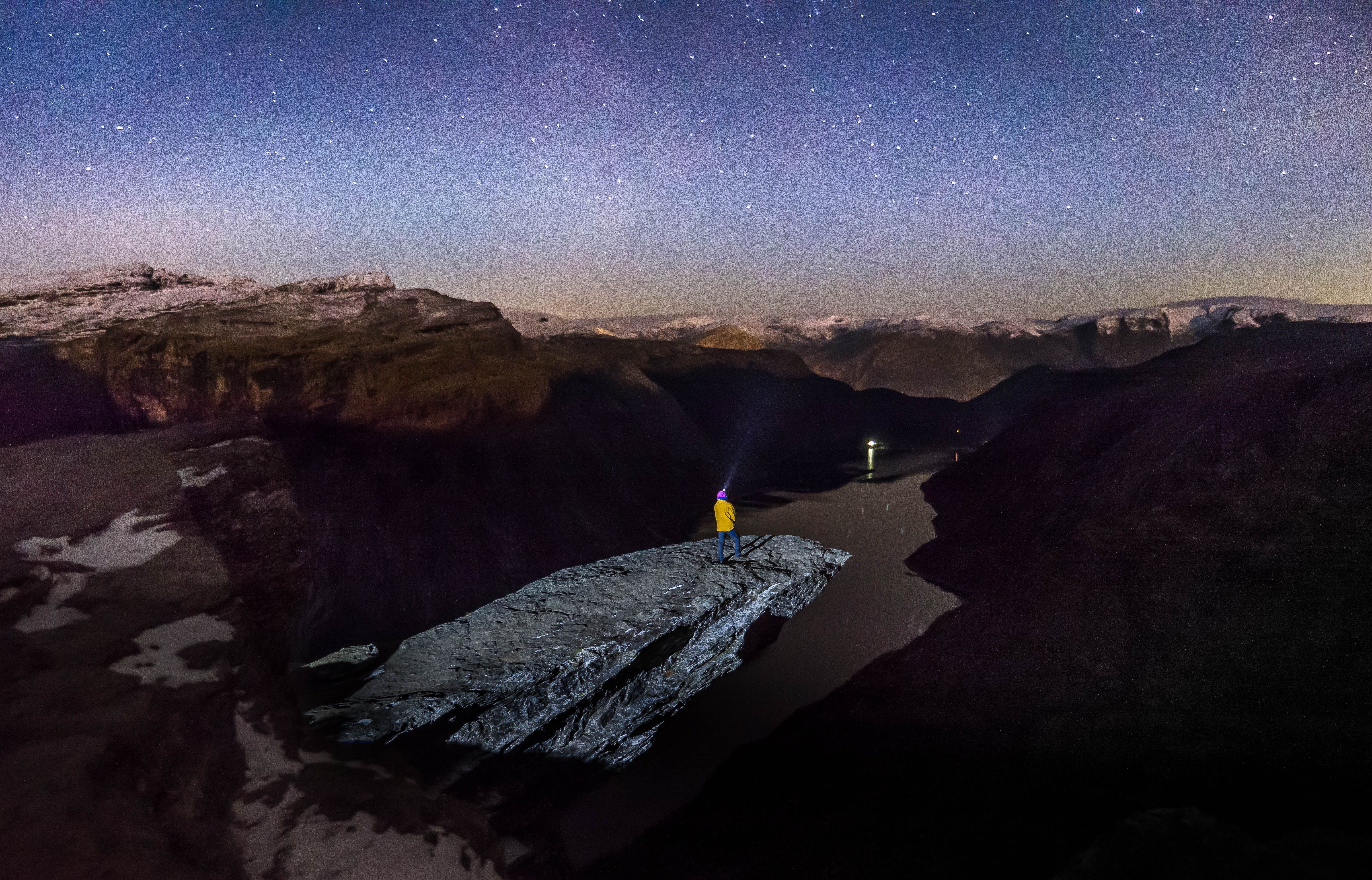 Hiker with headlamp standing on Trolltunga, overlooking the fjord under the night sky.