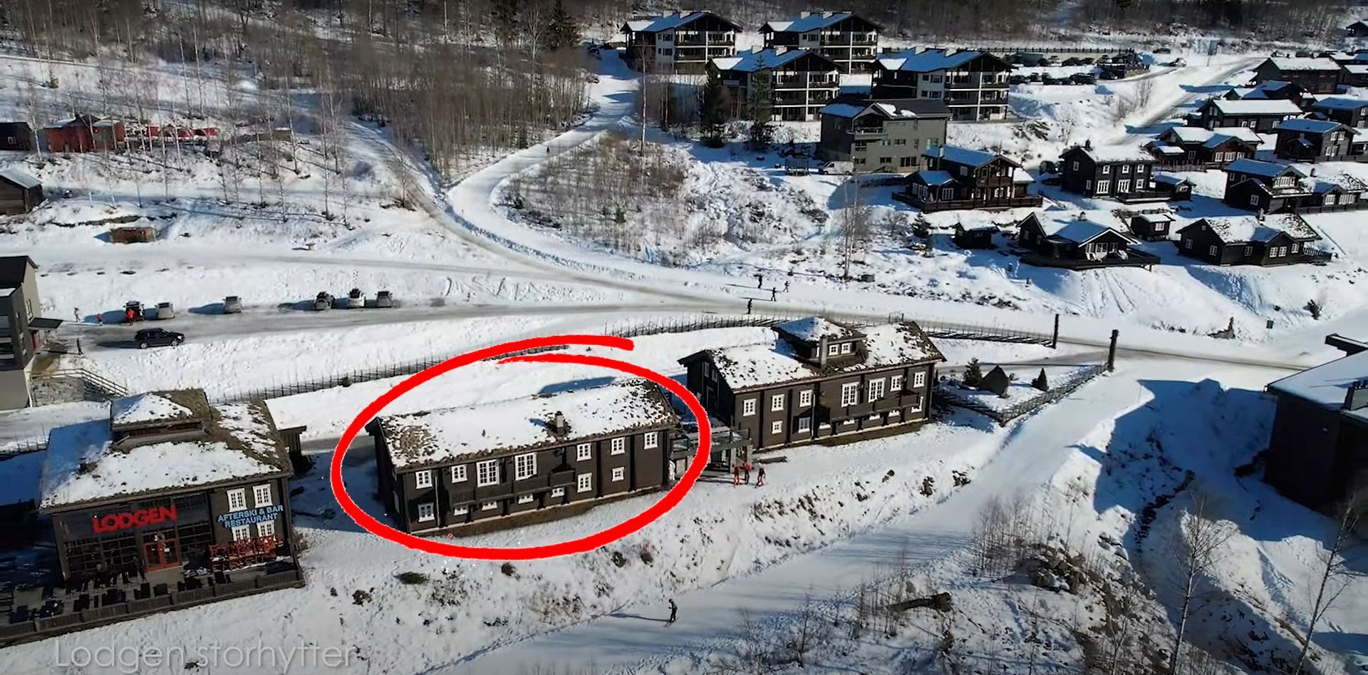 Flygfoto över en snötäckt fjällby med mörka trähus; en byggnad är markerad med en röd cirkel.