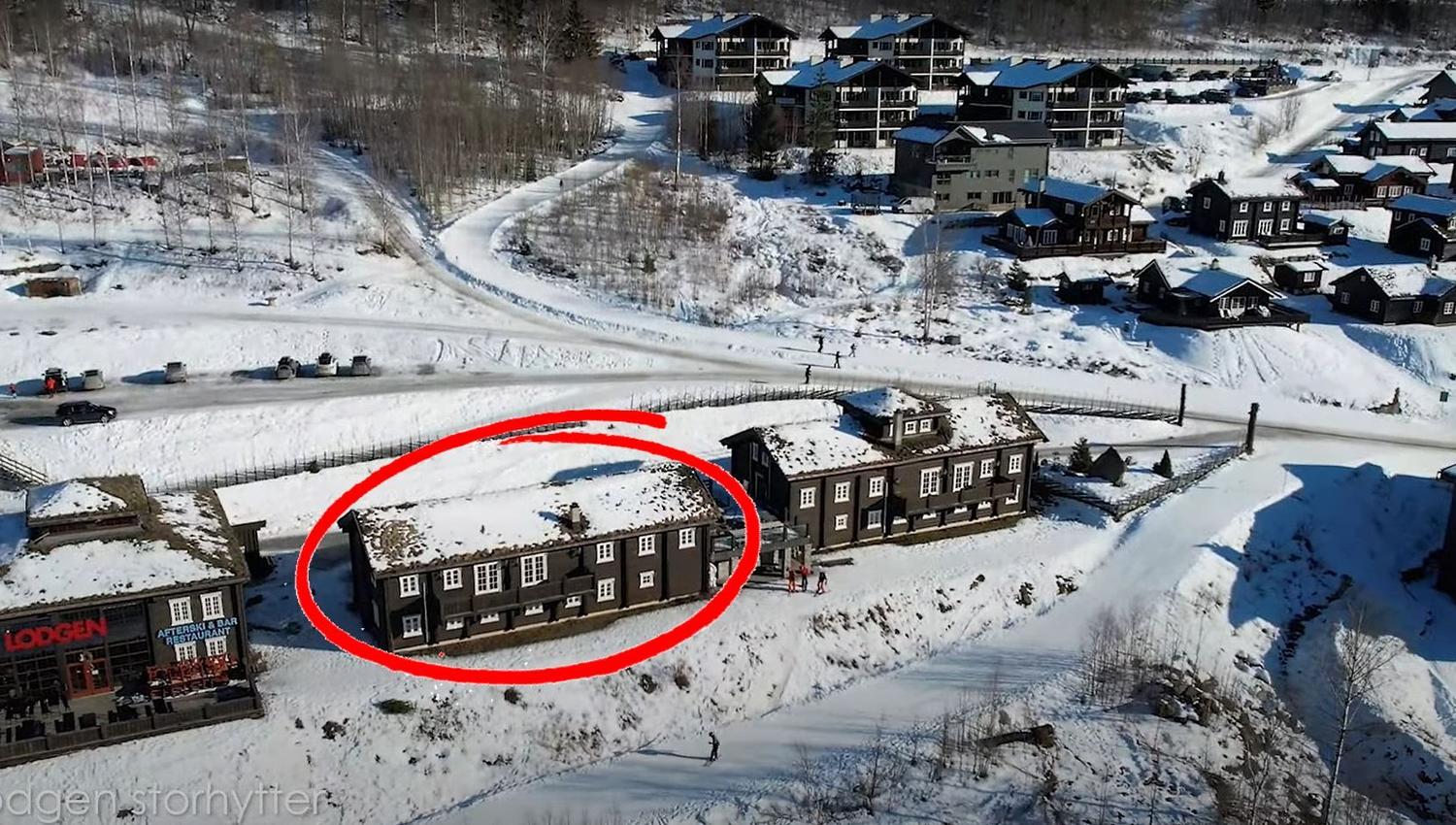 Flygfoto över en snötäckt fjällby med mörka trähus; en byggnad är markerad med en röd cirkel.
