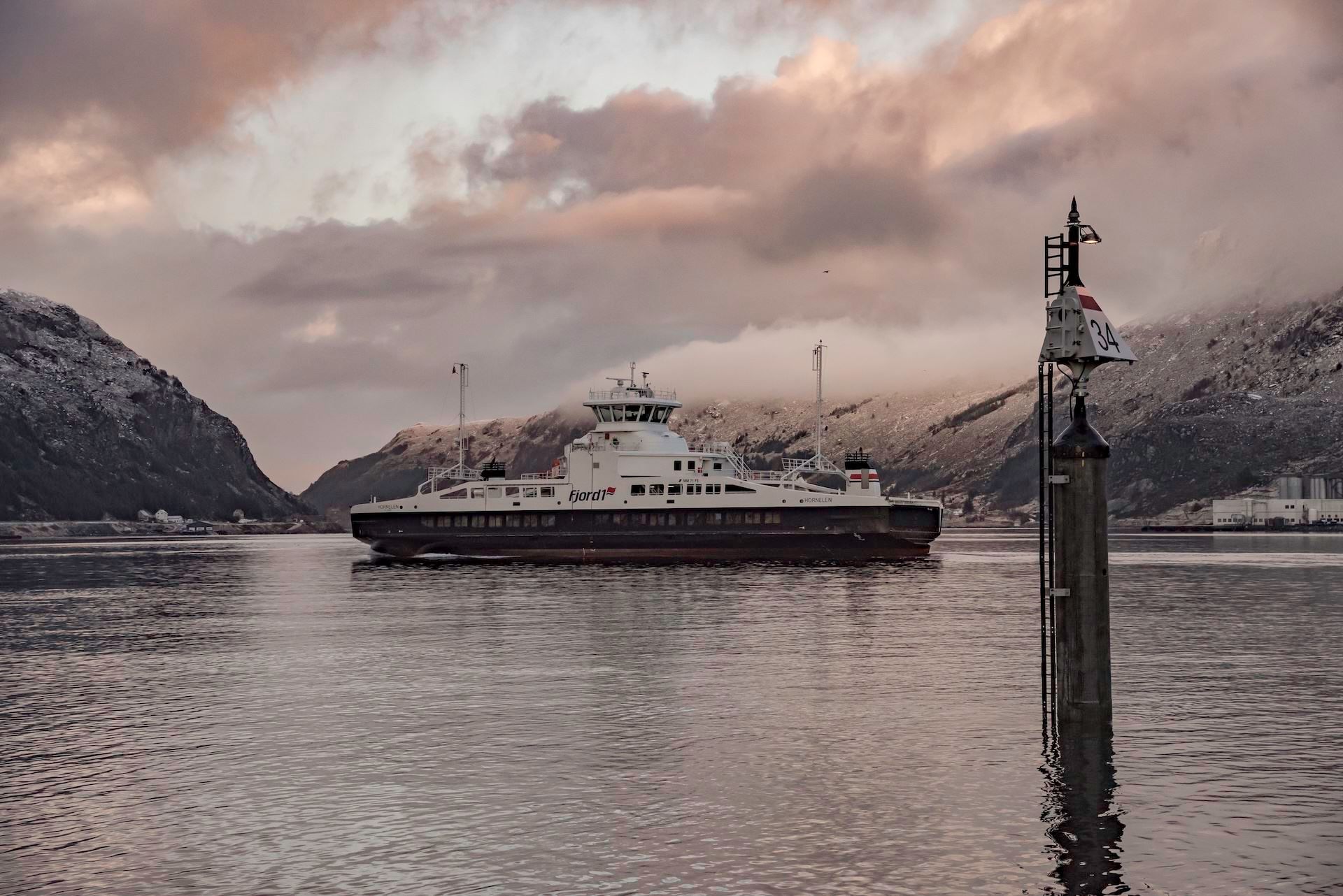 A car ferry travels across calm water with snowy mountains and pink clouds, beside a tall channel marker..