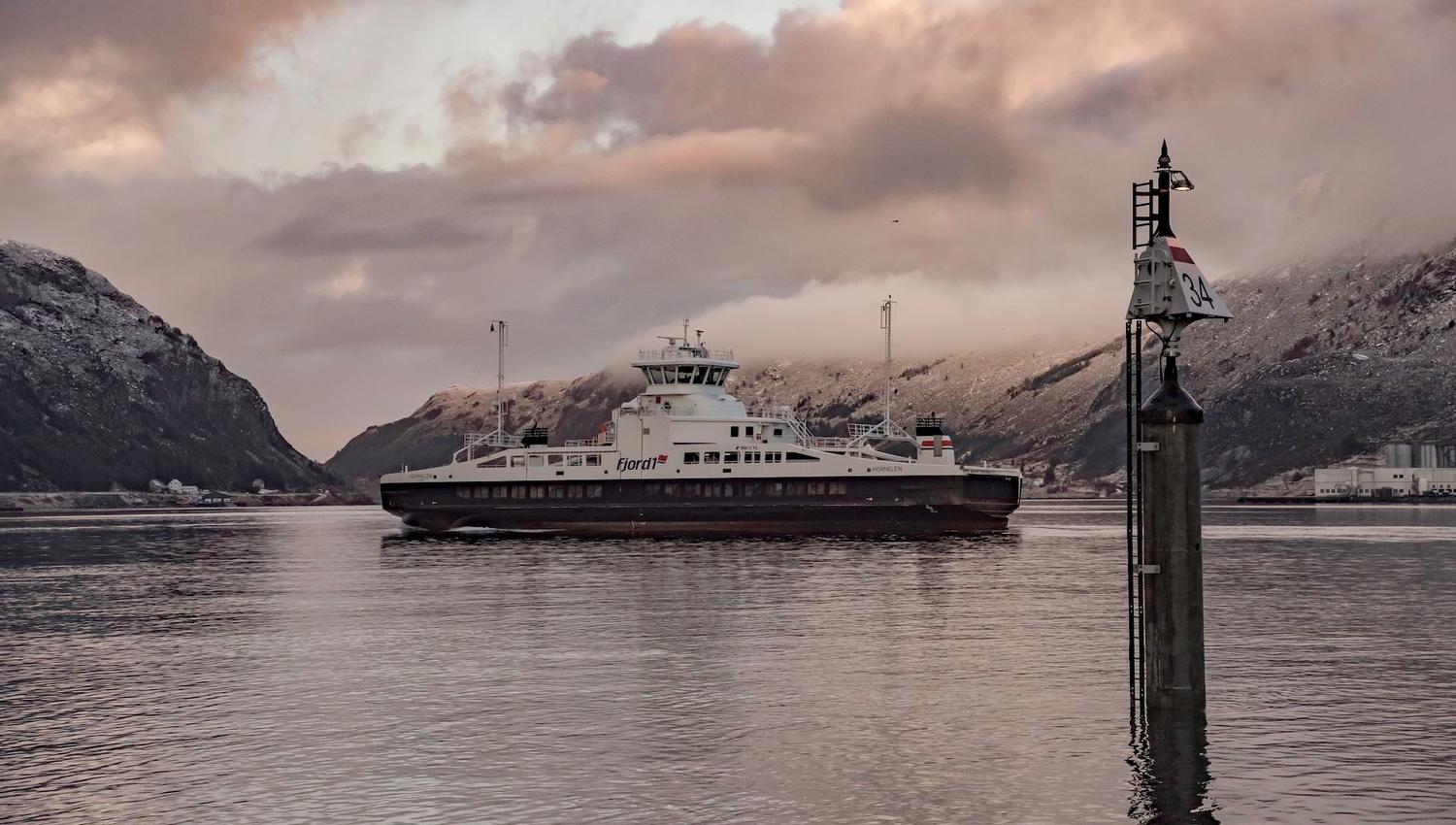 A car ferry travels across calm water with snowy mountains and pink clouds, beside a tall channel marker..