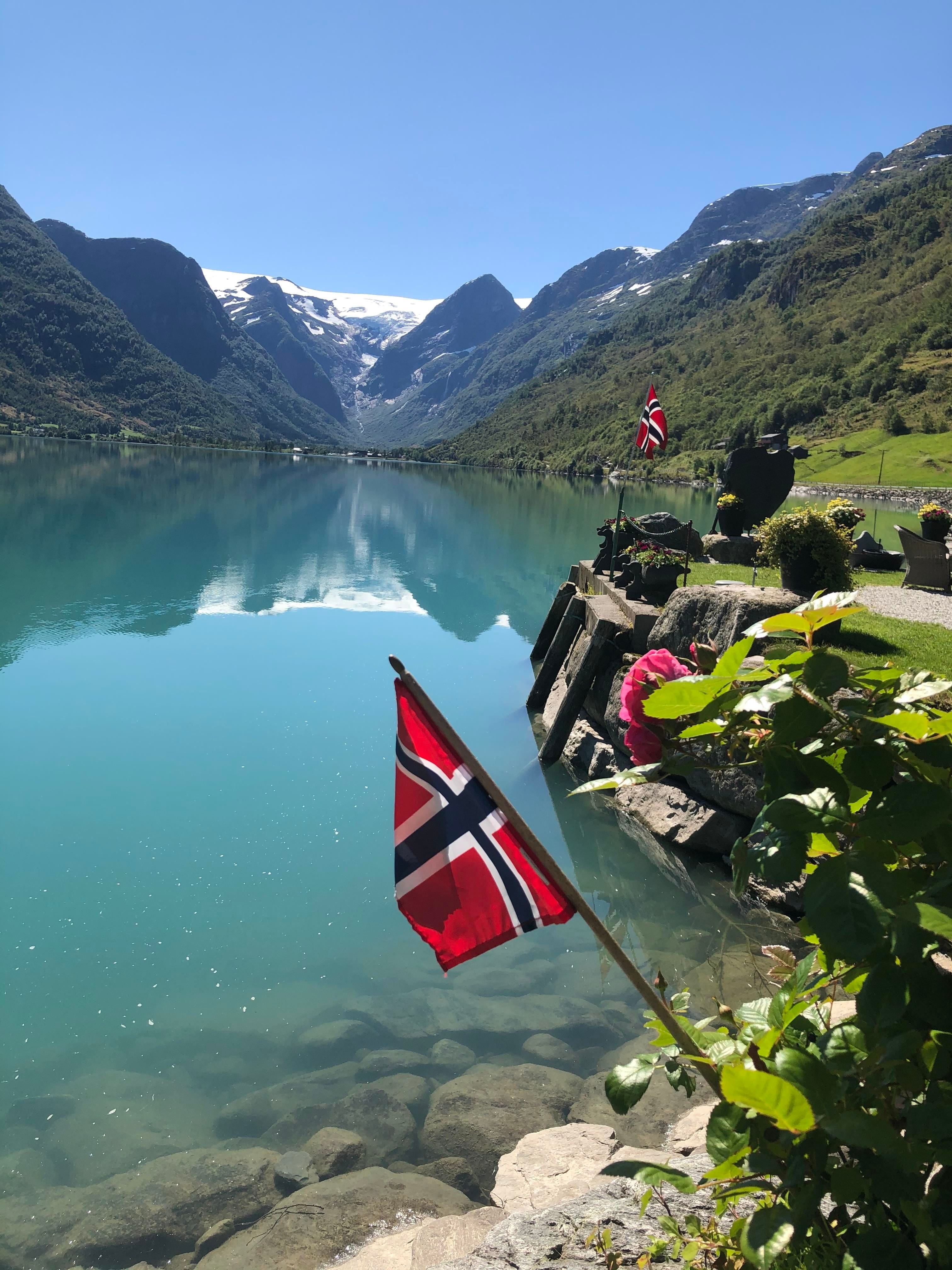 Norwegian flags by a rocky shoreline beside a calm turquoise fjord, with green mountains and snowy peaks under clear sky.
