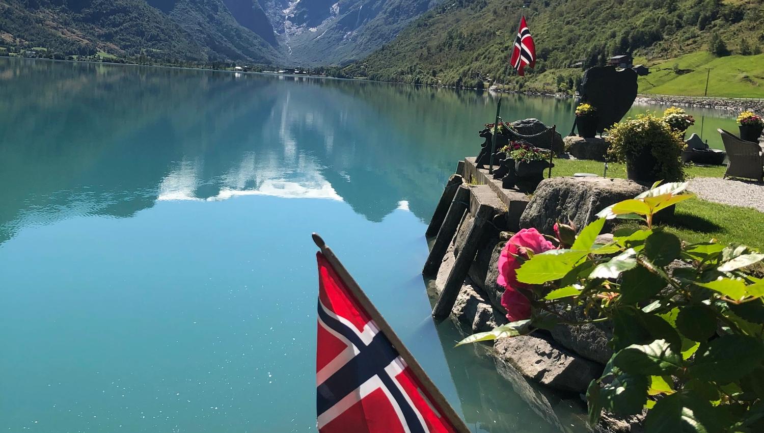 Norwegian flags by a rocky shoreline beside a calm turquoise fjord, with green mountains and snowy peaks under clear sky.