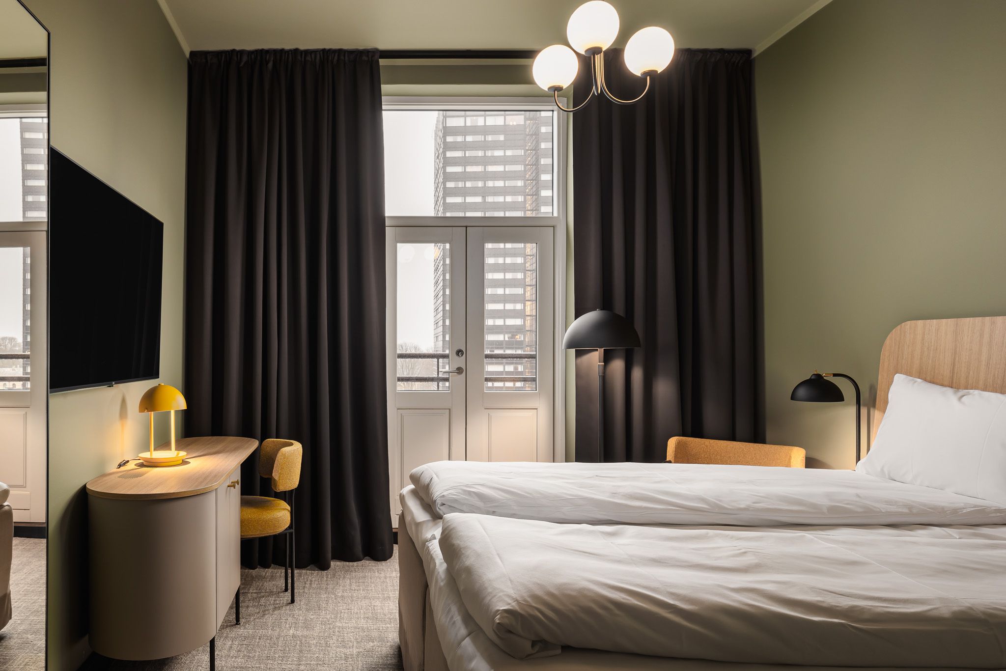 A hotel room with a double bed, green-painted walls, large black curtains along the big panoramic windows, and a TV on the wall