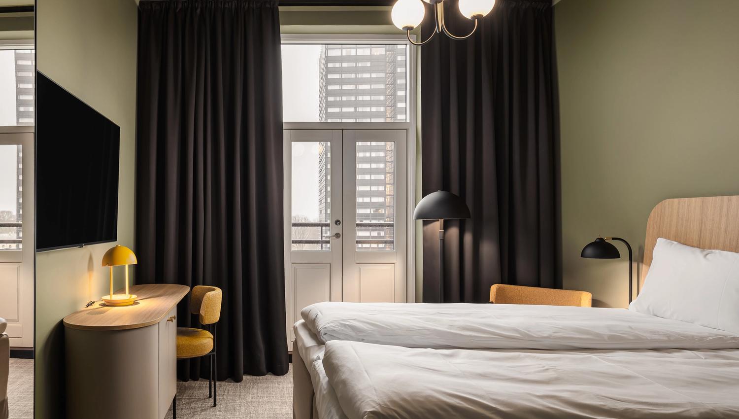 A hotel room with a double bed, green-painted walls, large black curtains along the big panoramic windows, and a TV on the wall
