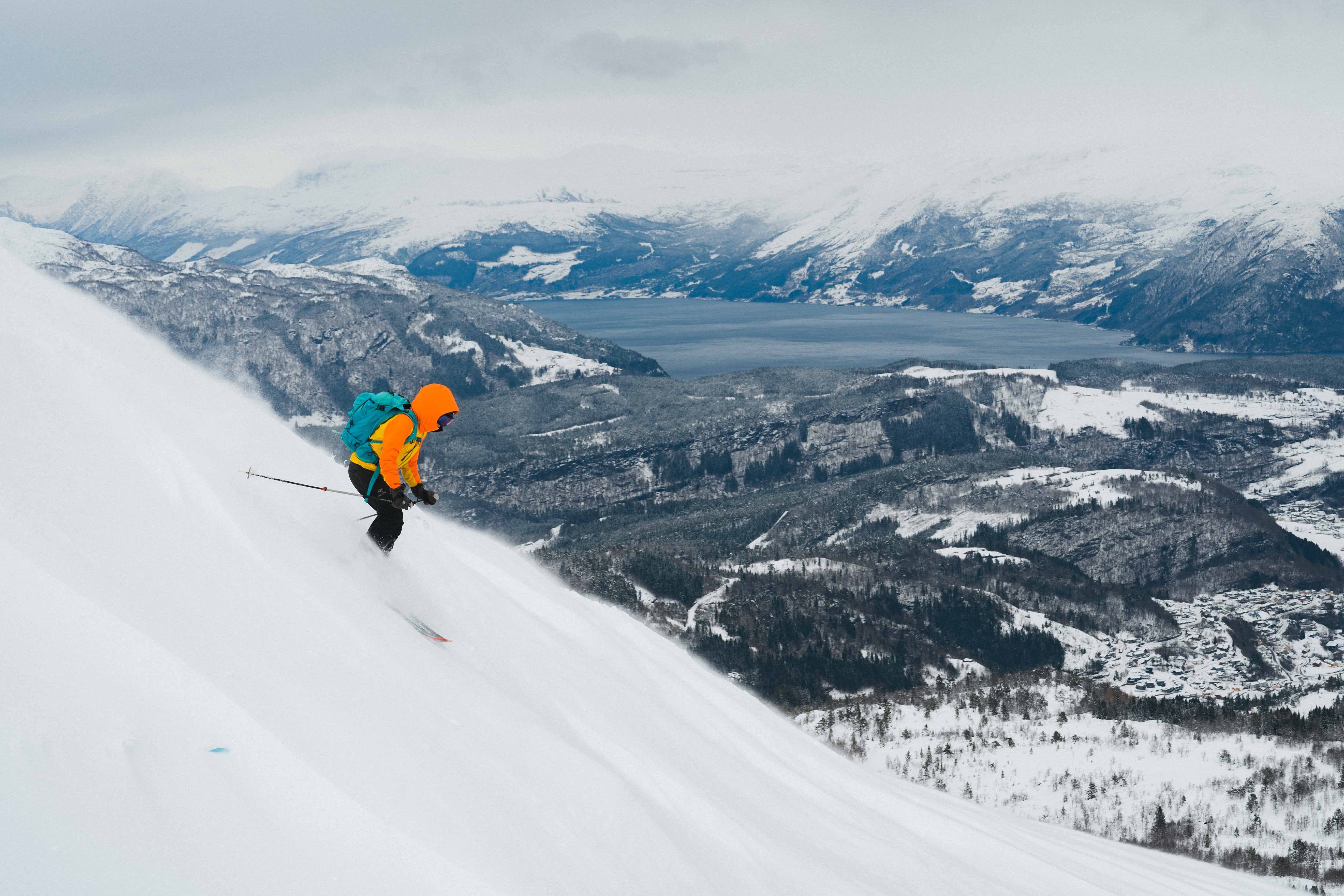 A person downhill skiing in a steep slope with views towards a fjord and snowy landscape. 