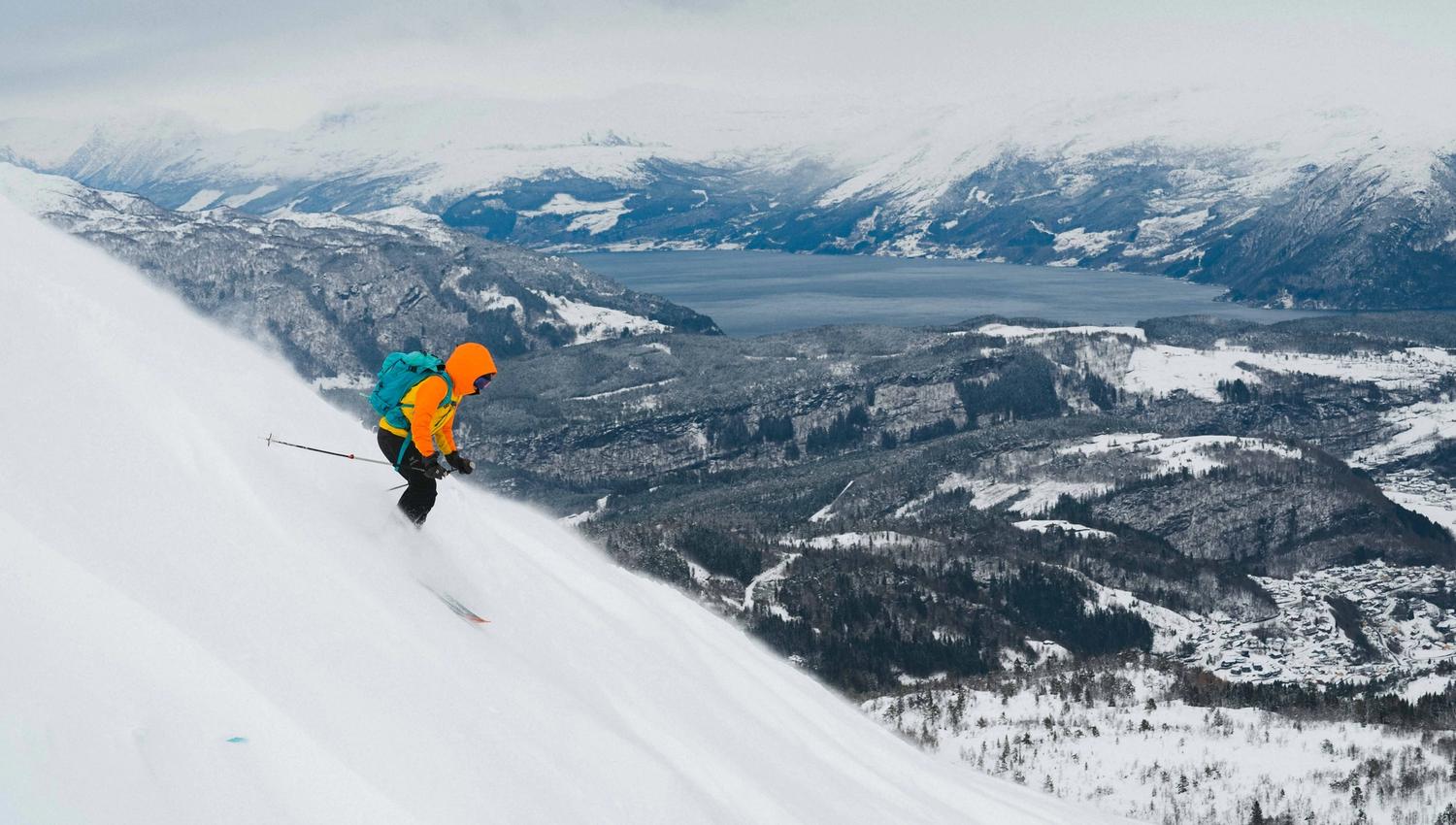 A person downhill skiing in a steep slope with views towards a fjord and snowy landscape.