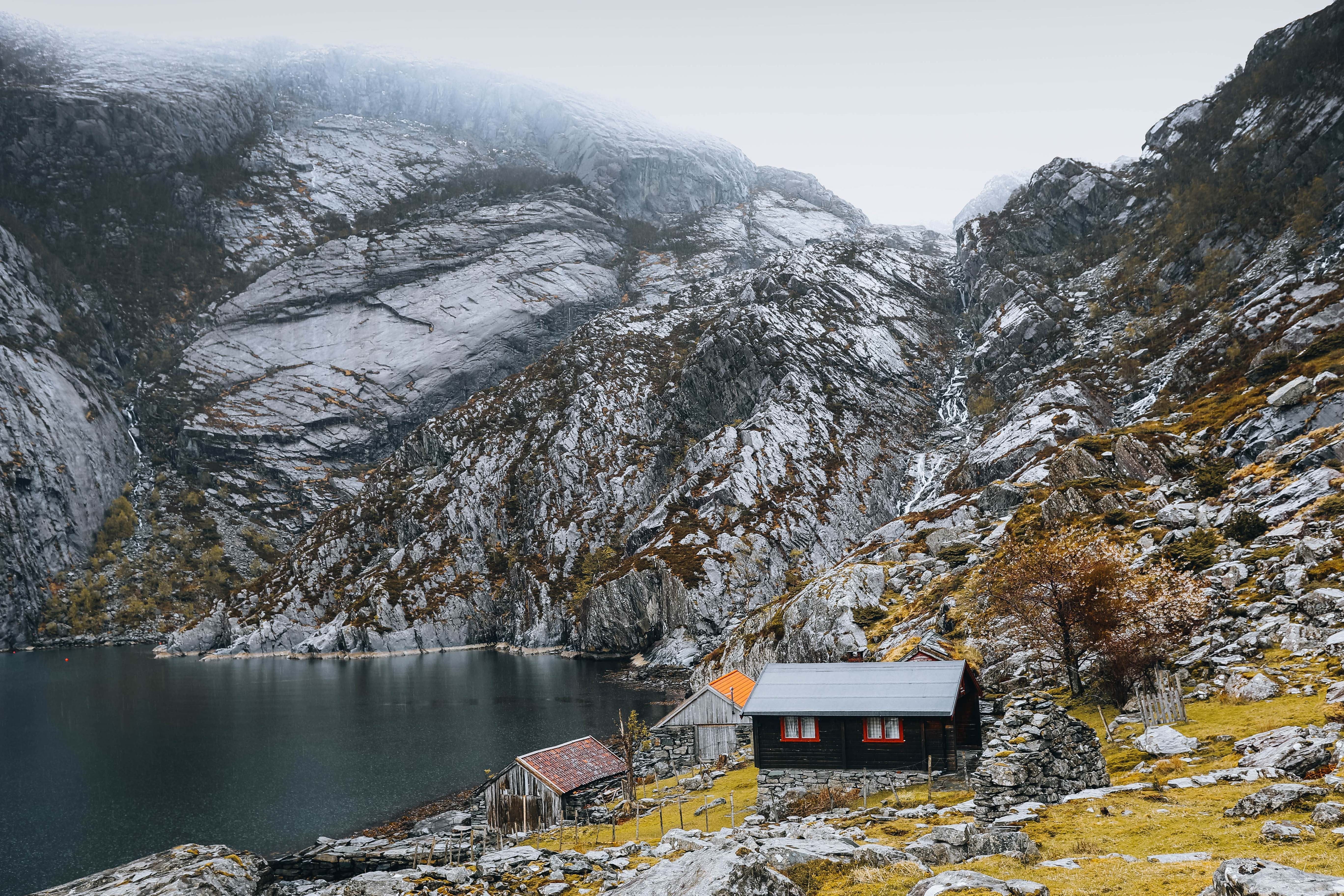 Small wooden cabins sit on a grassy shore beside a dark lake, backed by steep rocky mountains under misty skies.