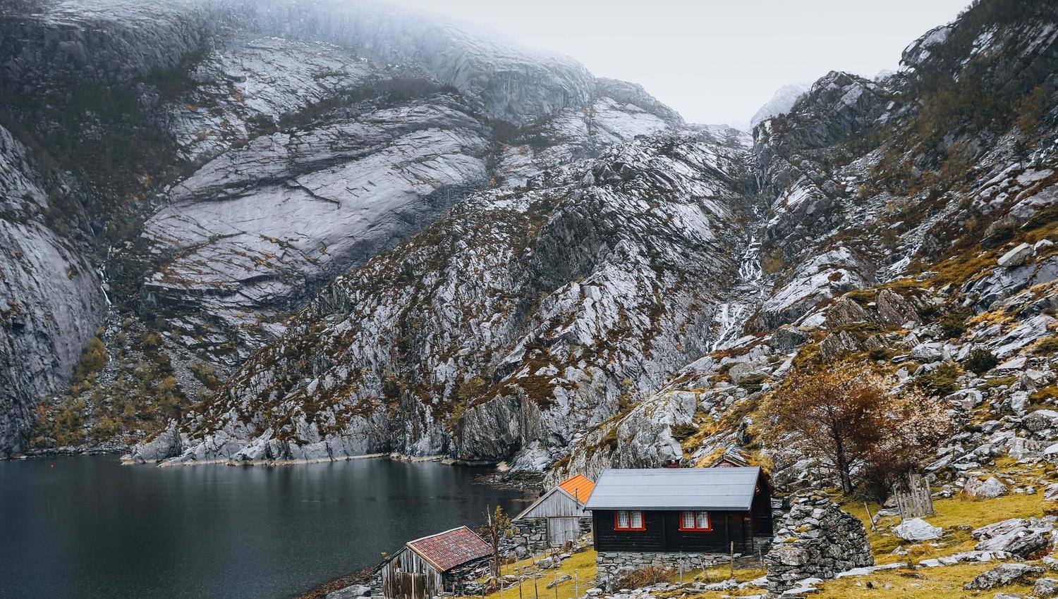 Small wooden cabins sit on a grassy shore beside a dark lake, backed by steep rocky mountains under misty skies.
