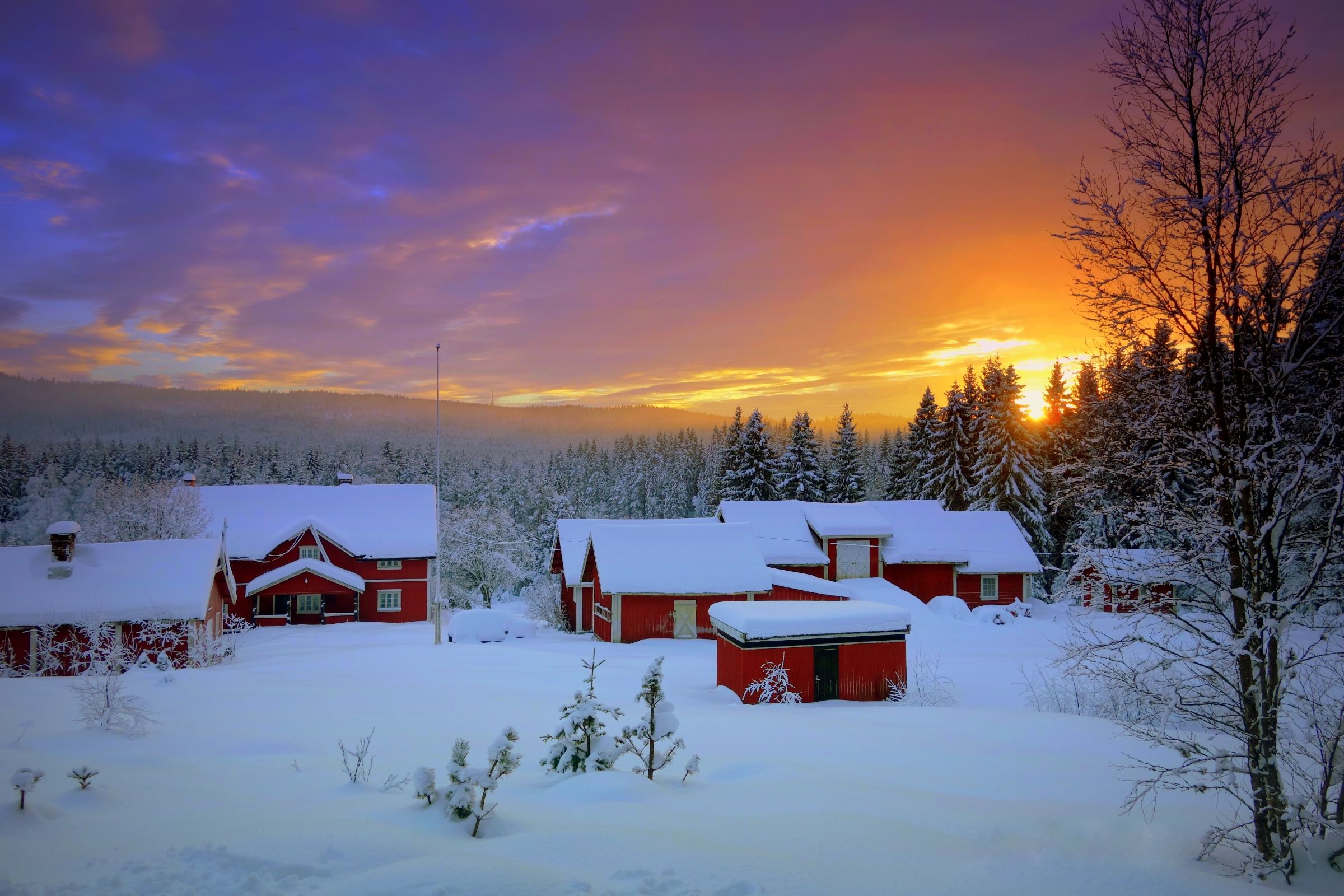 Wooden cottages covered in snow, with the sun setting in the background.