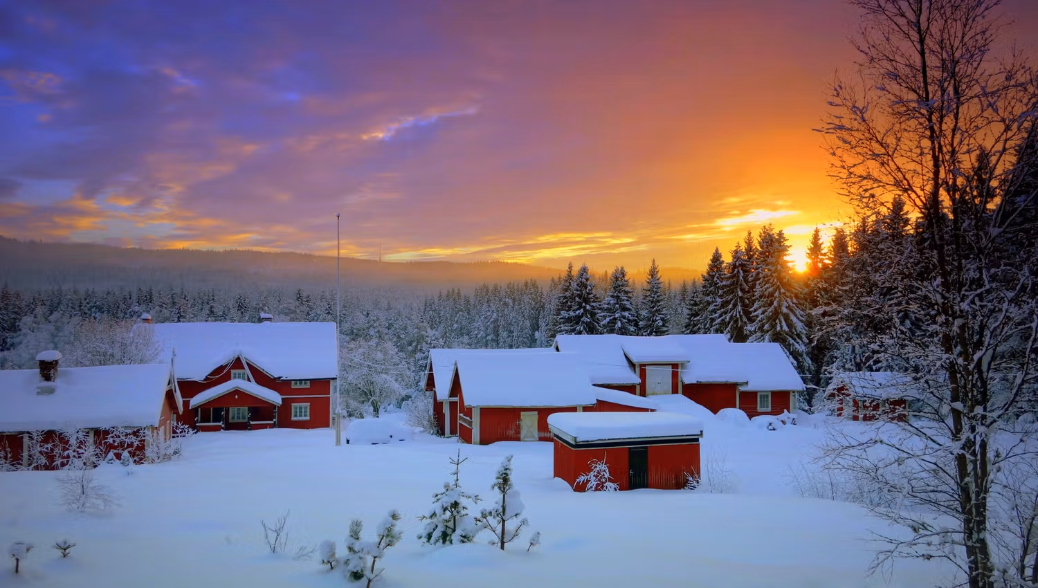 Wooden cottages covered in snow, with the sun setting in the background.