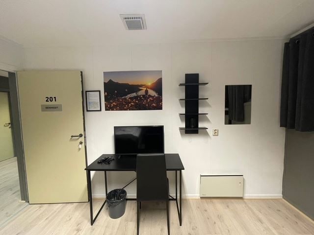 Small room with an open door, desk and chair facing a TV, wall shelves, framed landscape photo, and dark curtain.
