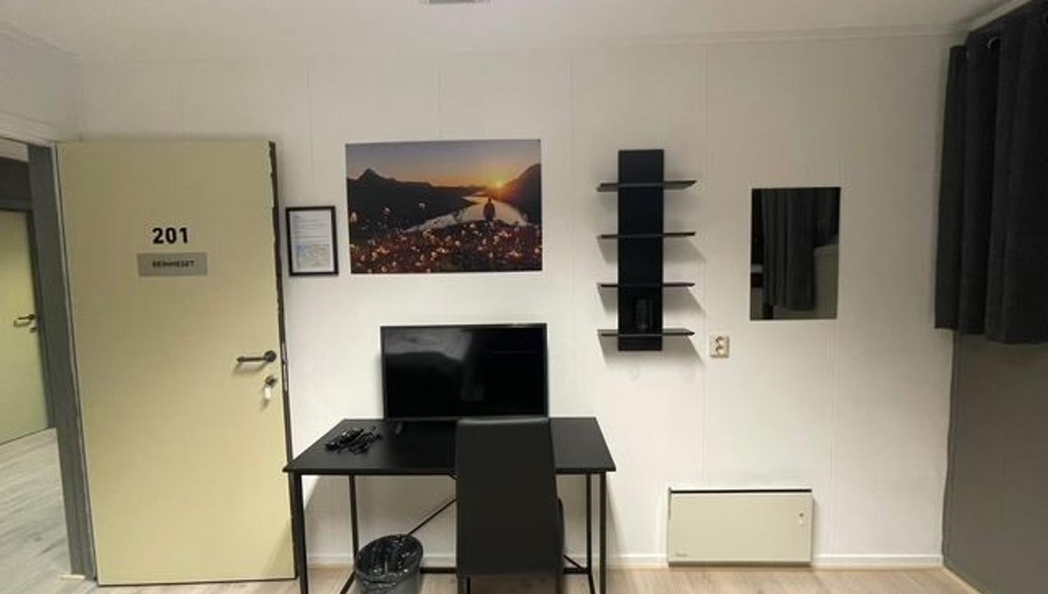 Small room with an open door, desk and chair facing a TV, wall shelves, framed landscape photo, and dark curtain.