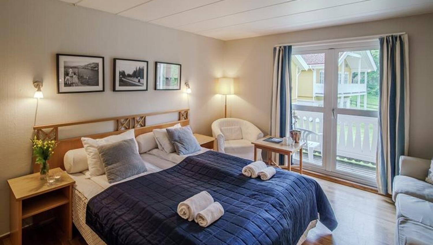 Lyst hotellrom på Hankø Hotell & Spa, med dobbeltseng, blått sengeteppe, sittegruppe og balkongdør med utsikt mot hagen.
