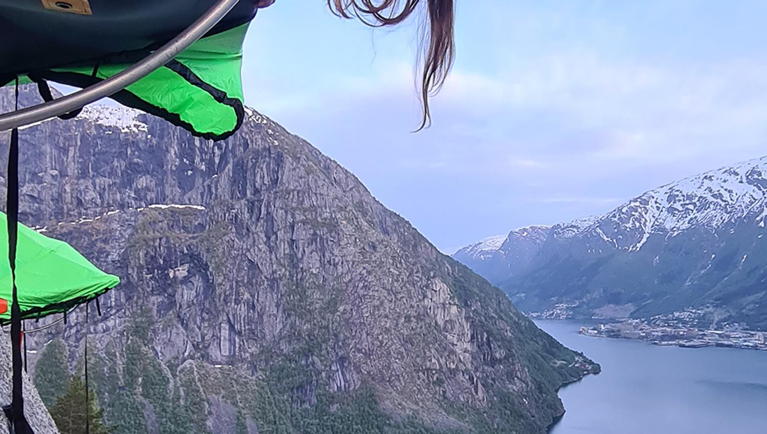 Ei kvinne smilar og stikk hovudet ut frå ein portaledge, med fjorden langt under ho i Hardanger. Eit eventyrleg overnattingsalternativ.