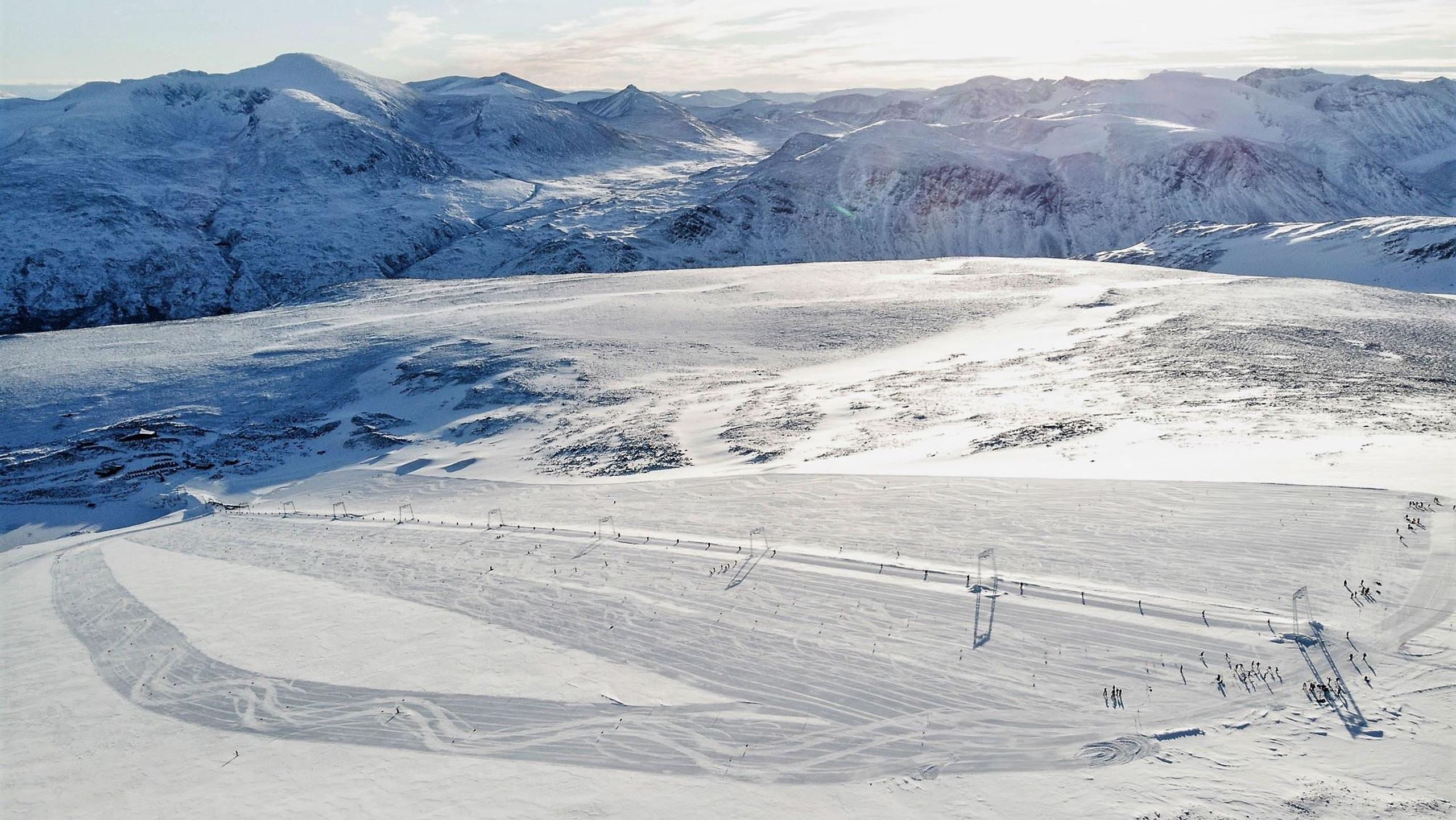 Wide view of snow-covered mountains and a groomed ski area with scattered skiers under bright winter sunlight..