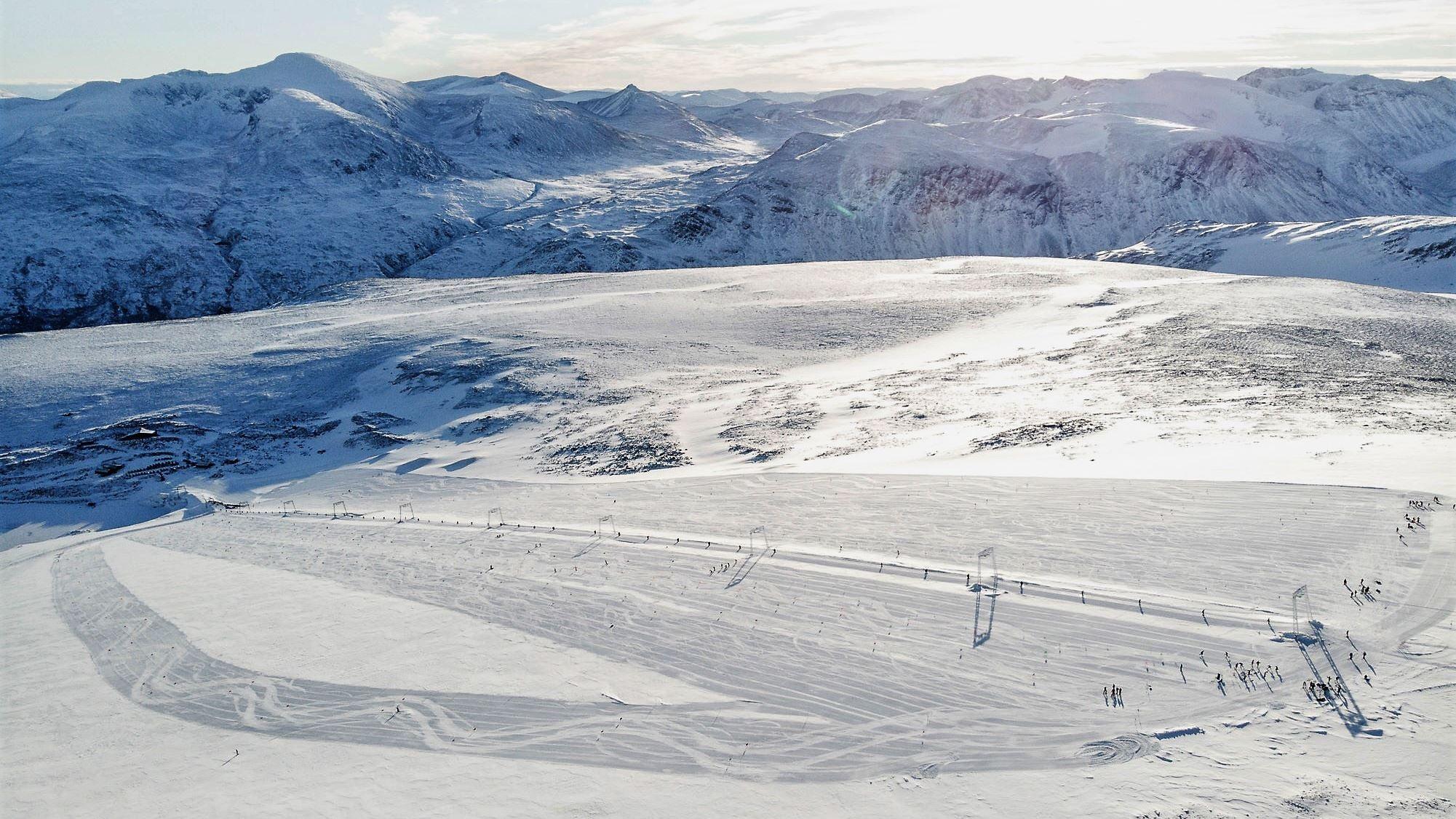 Wide view of snow-covered mountains and a groomed ski area with scattered skiers under bright winter sunlight..