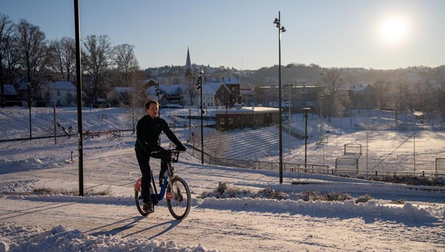 Mann som sykler på en snødekt vei, med en fotballbane i bakgrunnen
