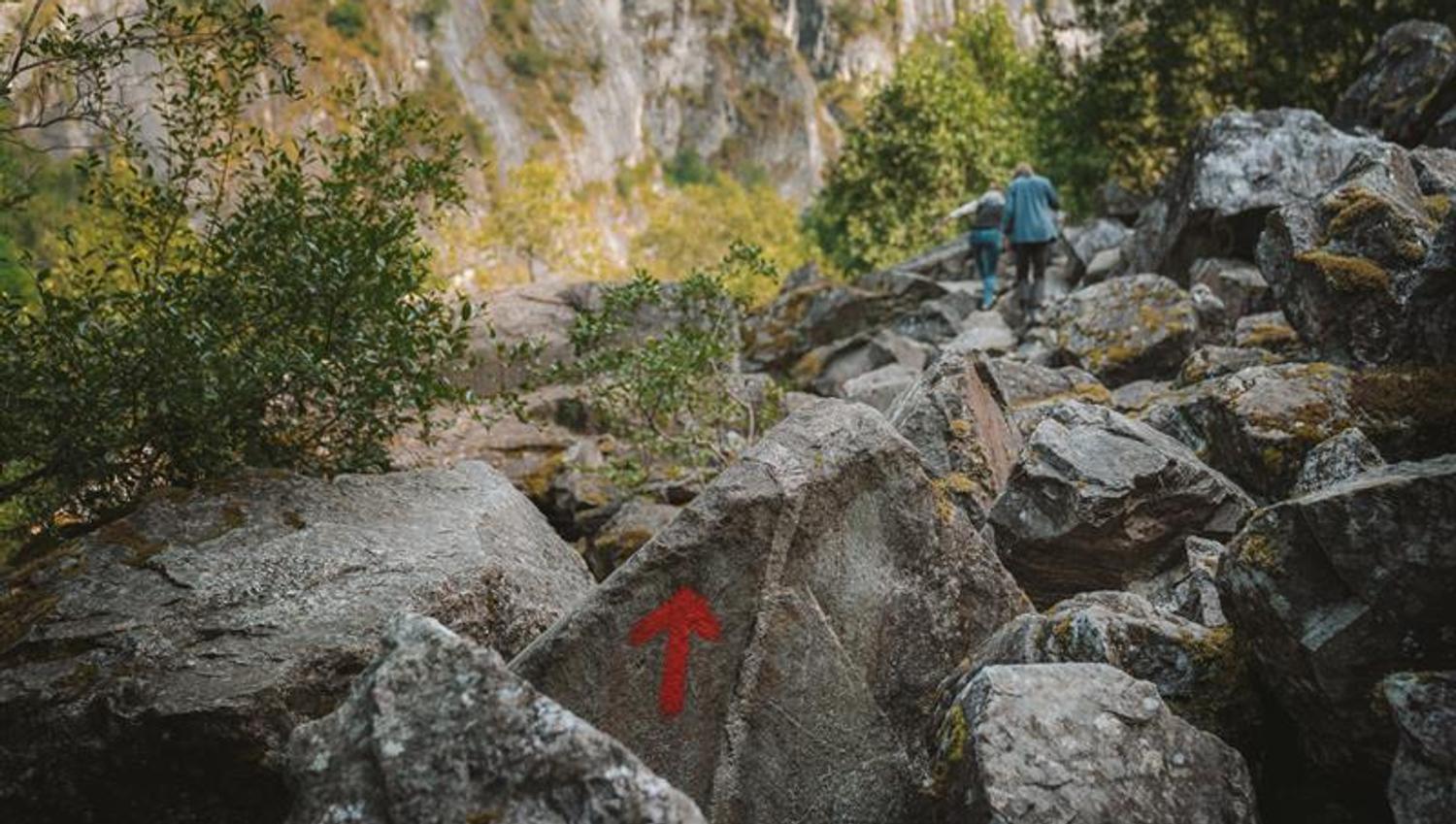 A large rock painted with a red arrow with a couple of hikers seen far in the background.