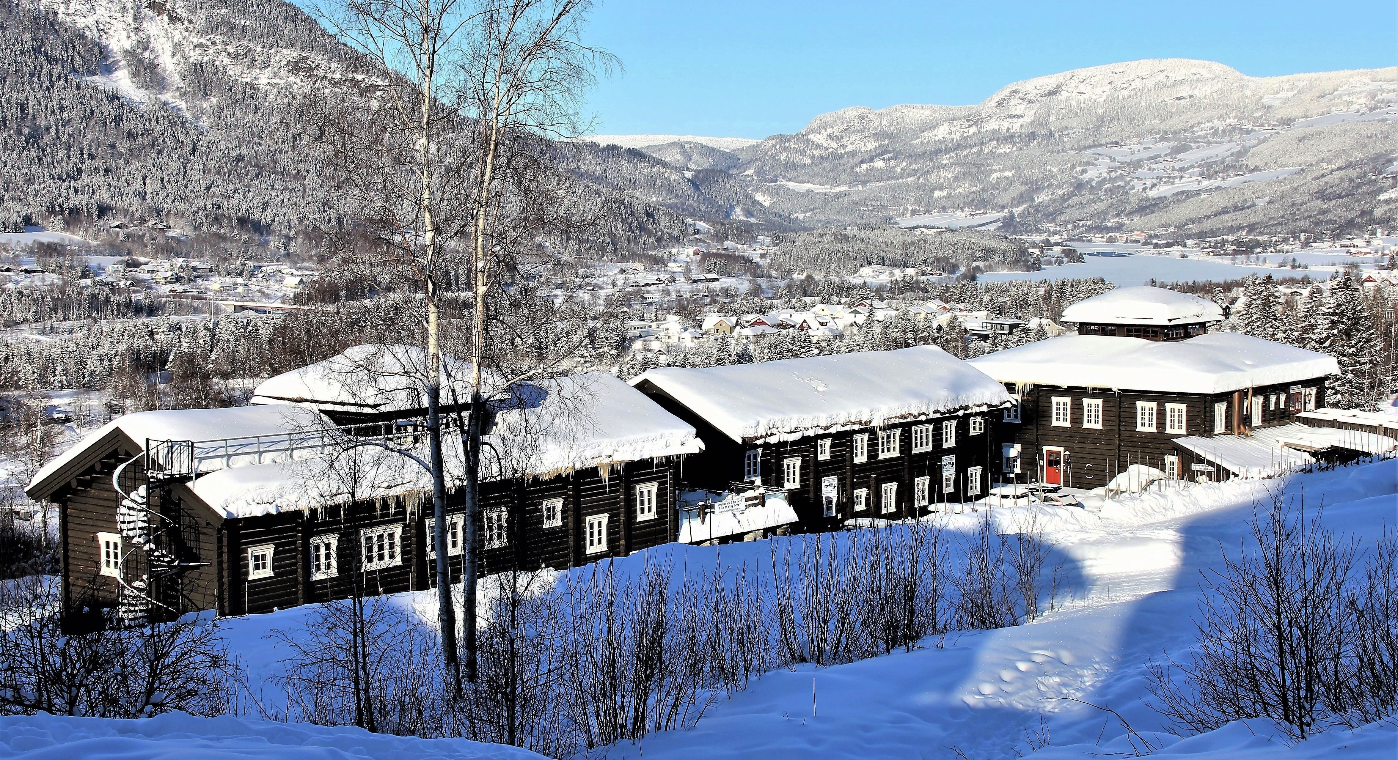 Panoramavy över en snötäckt dal med mörka träbyggnader i förgrunden, fjäll och by i bakgrunden.
