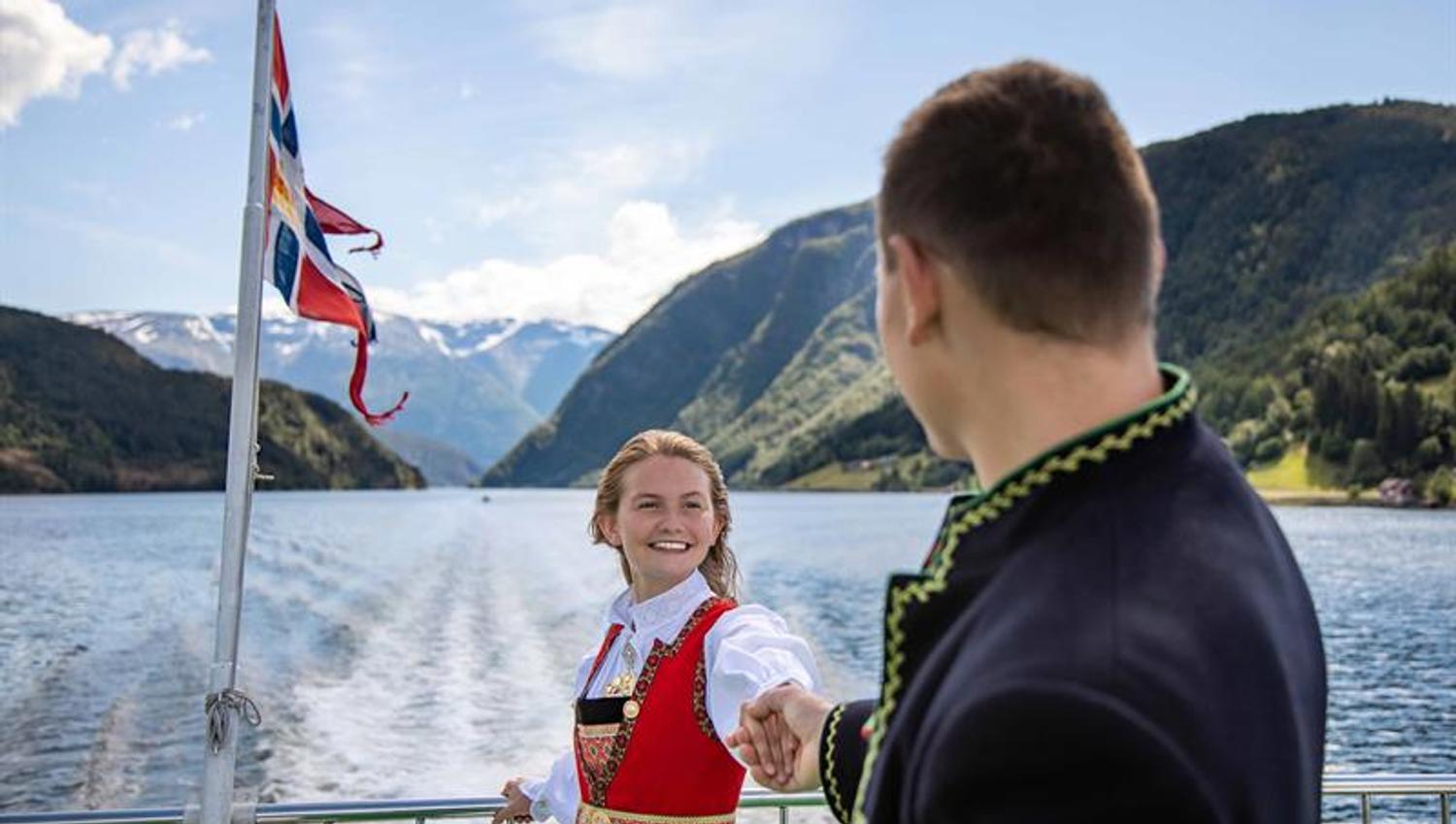 Par i bunad held kvarandre i handa på akterdekket under fjordcruise på Hardangerfjorden, norsk flagg i bakgrunnen.