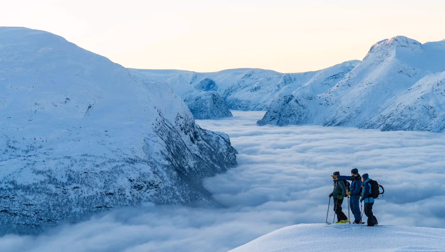 Three hikers stand on a snowy ridge overlooking cloud-filled valleys and snow-covered mountains at sunrise.
