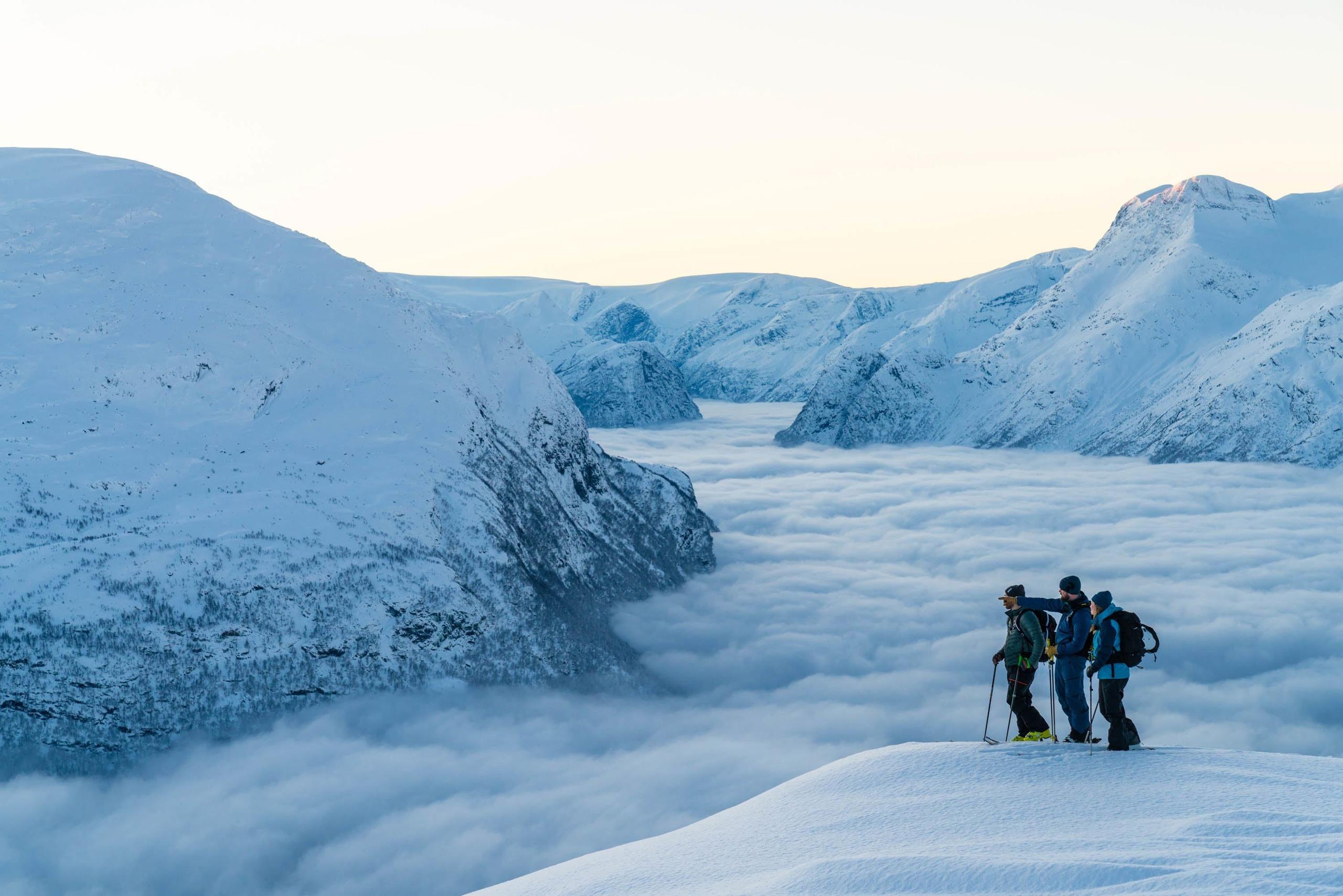 Three hikers stand on a snowy ridge overlooking cloud-filled valleys and snow-covered mountains at sunrise.