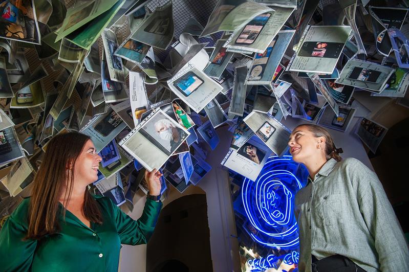 Two people inside an installation featuring suspended photographs, documents and illuminated artworks.
