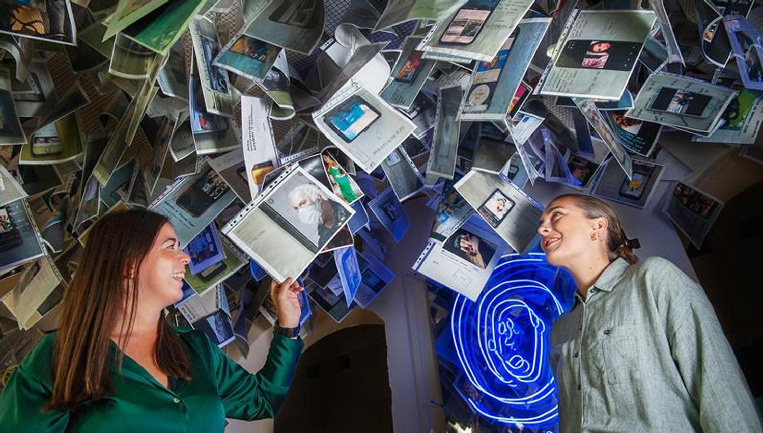 Two people inside an installation featuring suspended photographs, documents and illuminated artworks.
