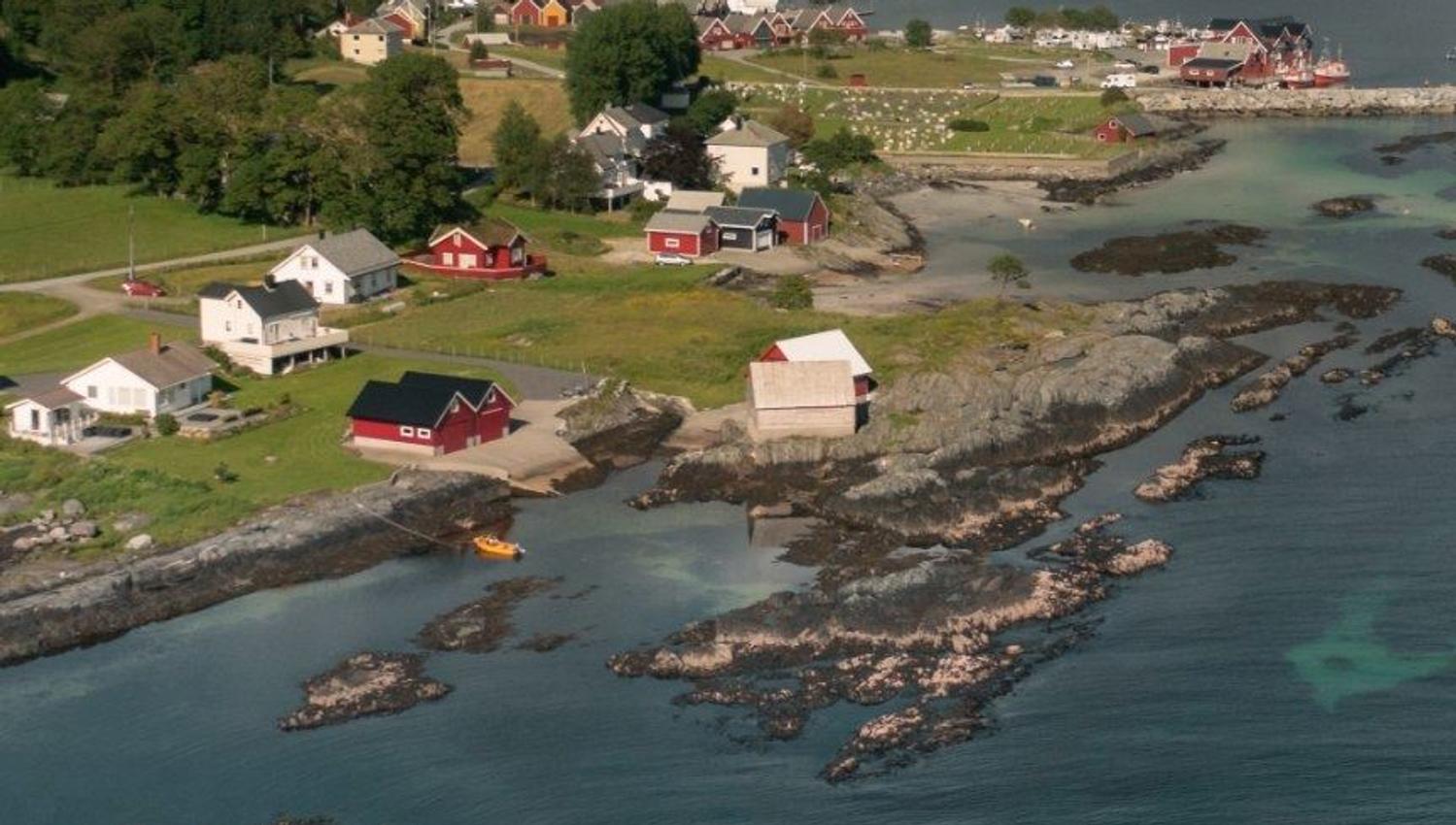 Aerial view of a coastal village with scattered houses, rocky shoreline, green hills, and a boat on turquoise water..