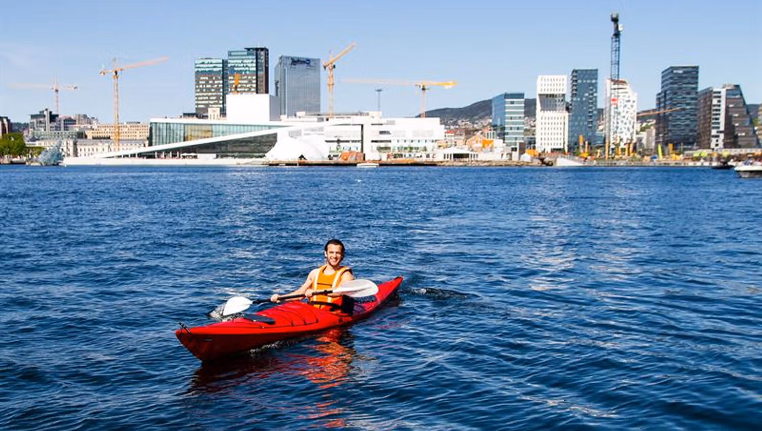 A man sitting in a red kayak in front of the Opera House and Barcode.