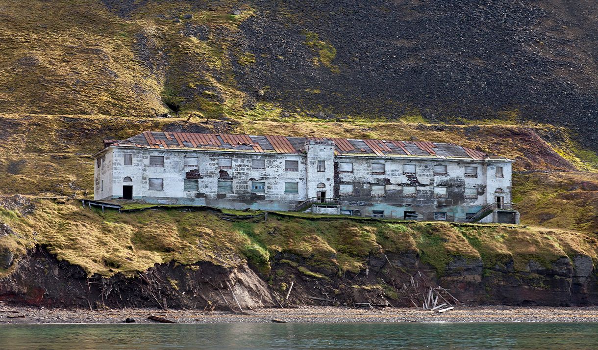En forlatt bygning i det gamle gruvesamfunnet Grumant, med en strand og fjord i forgrunnen og en fjellside i bakgrunnen