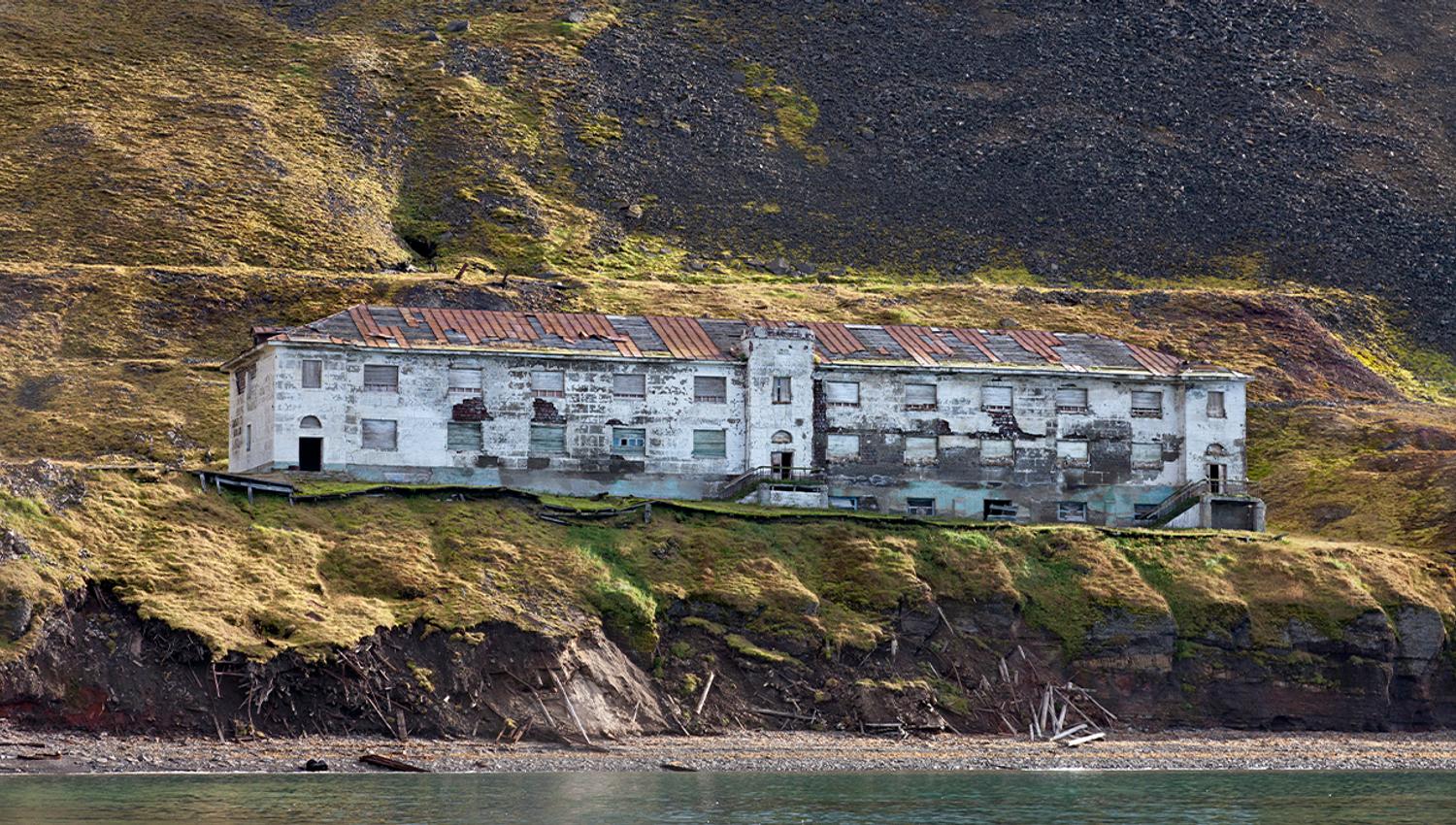En forlatt bygning i det gamle gruvesamfunnet Grumant, med en strand og fjord i forgrunnen og en fjellside i bakgrunnen