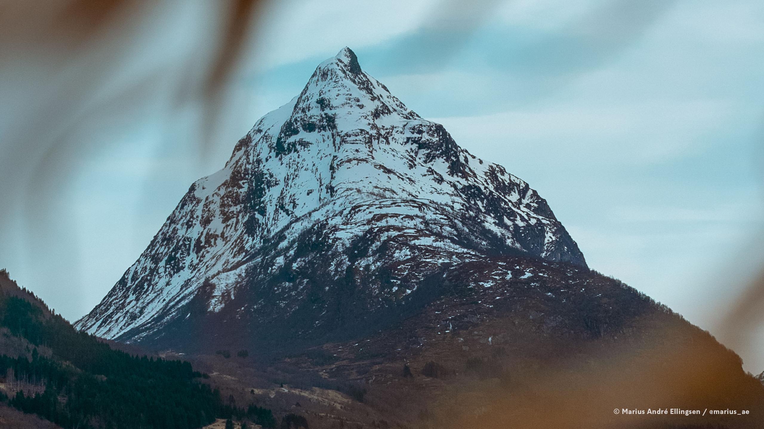 Snødekt, spiss fjelltopp med mørke fjellsider og skogkledde skråningar under ein lys, blå himmel..