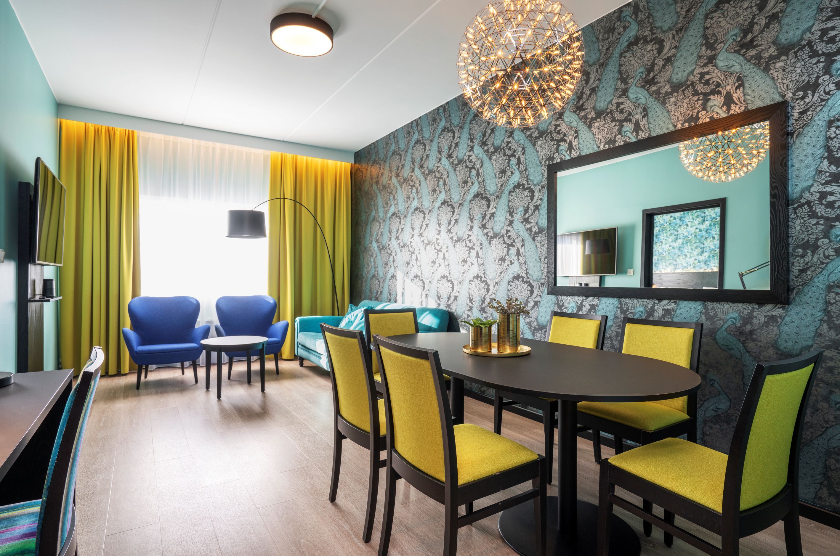 Hotellrom med mønstret tapet, sort spisebord og gule stoler, blå, sofa, blå lenestoler og gule gardiner