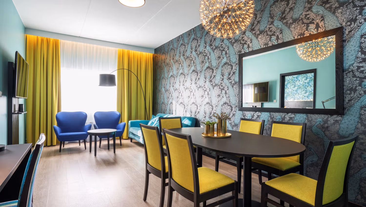 Hotellrom med mønstret tapet, sort spisebord og gule stoler, blå, sofa, blå lenestoler og gule gardiner