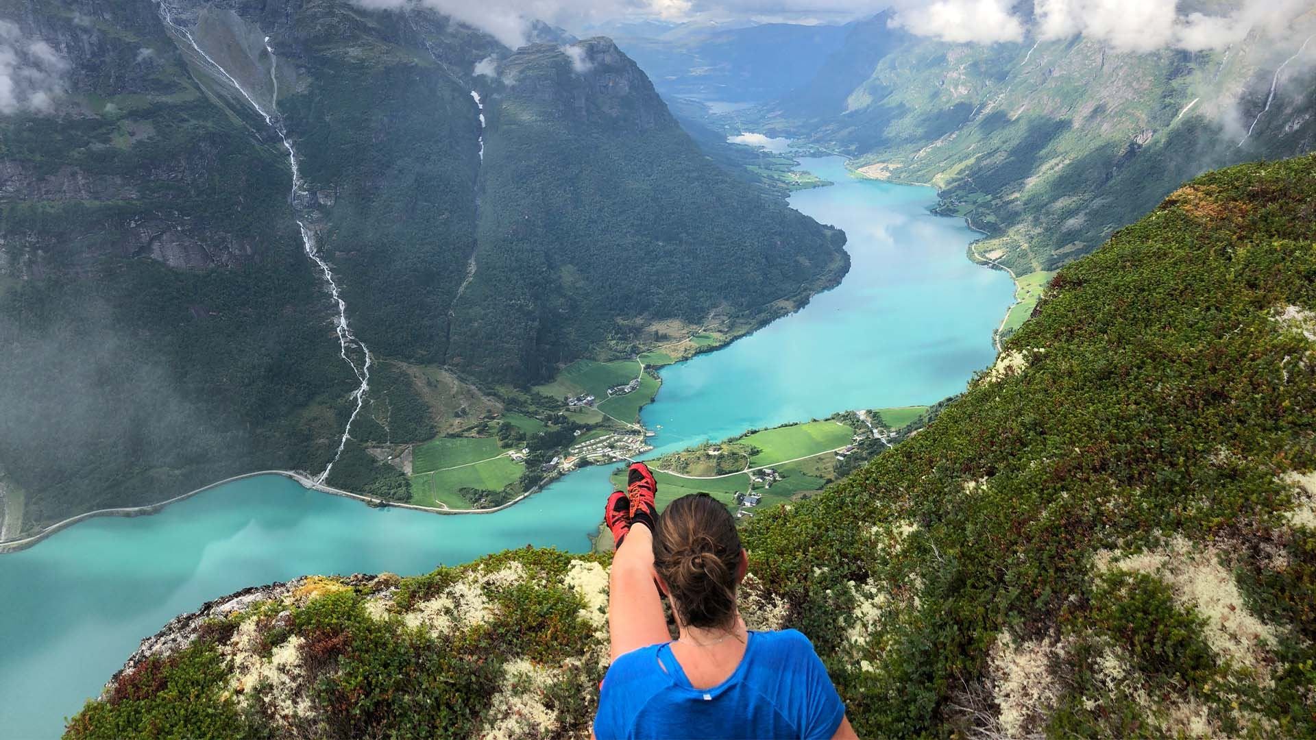 A hiker sits on a grassy cliff, looking over a turquoise fjord, green valleys, steep mountains, and clouds..