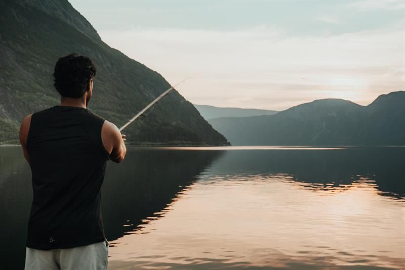 A man is fishing by the fjord during sunset.