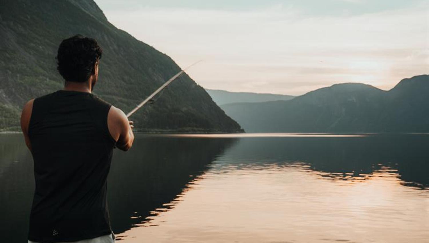 A man is fishing by the fjord during sunset.
