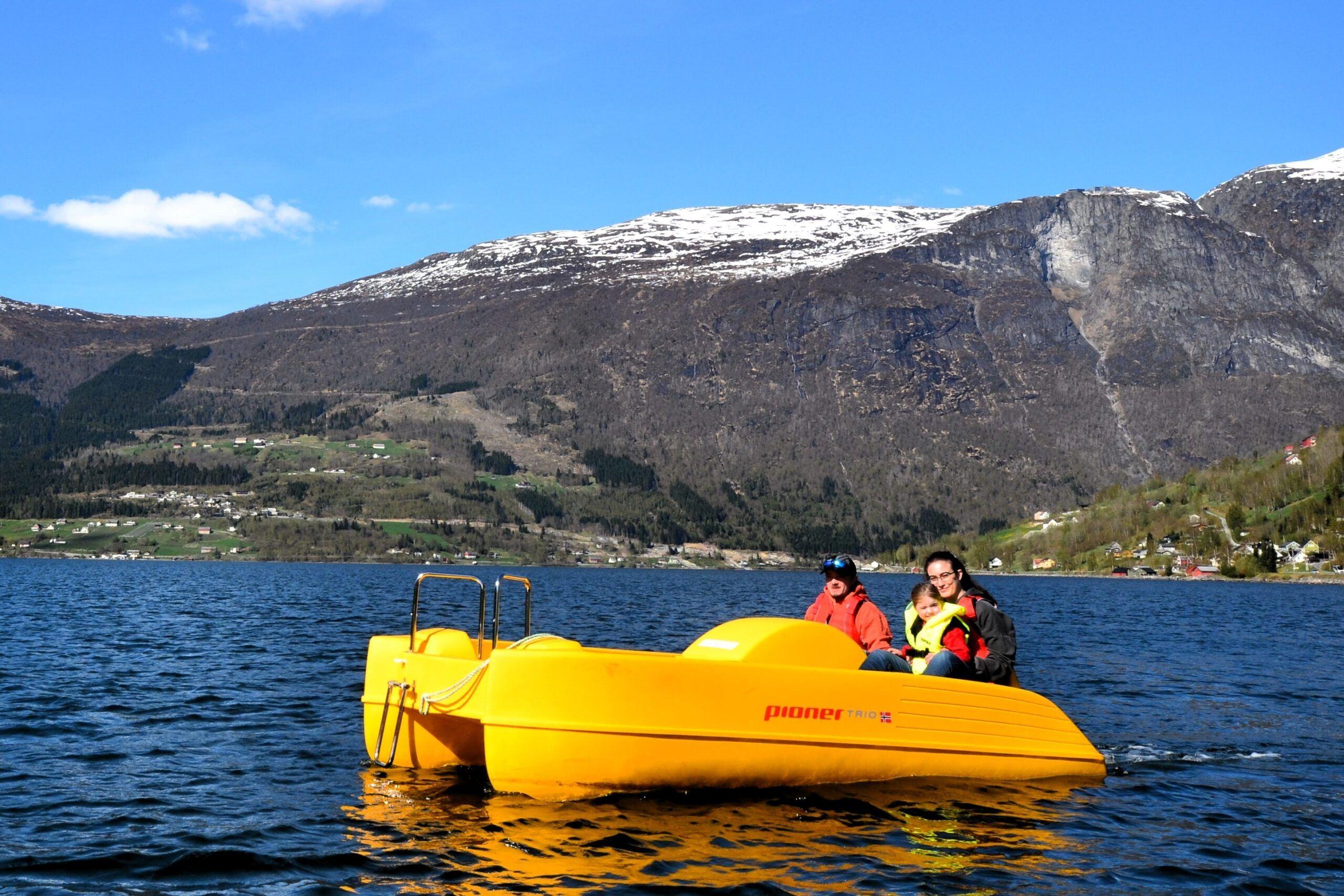 Three people ride a bright yellow pedal boat on a blue Nordfjord fjord, with snow-dusted mountains and a clear sky behind in Olden..
