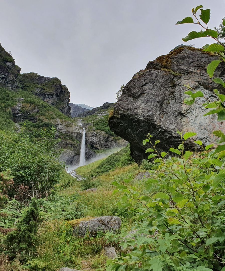 A tall waterfall cascades between rocky cliffs into a lush green valley with shrubs, boulders, and overcast sky.