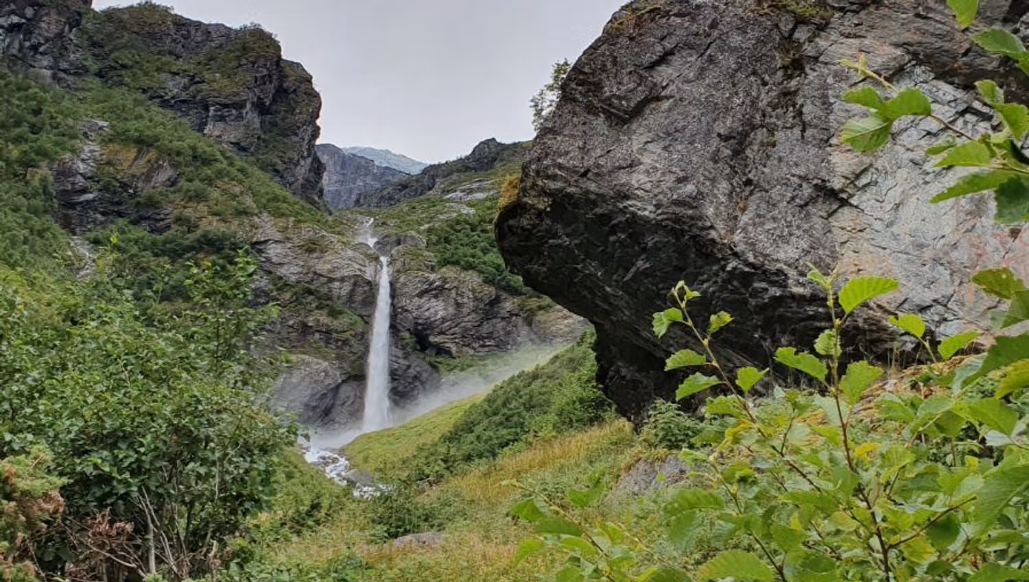 A tall waterfall cascades between rocky cliffs into a lush green valley with shrubs, boulders, and overcast sky.