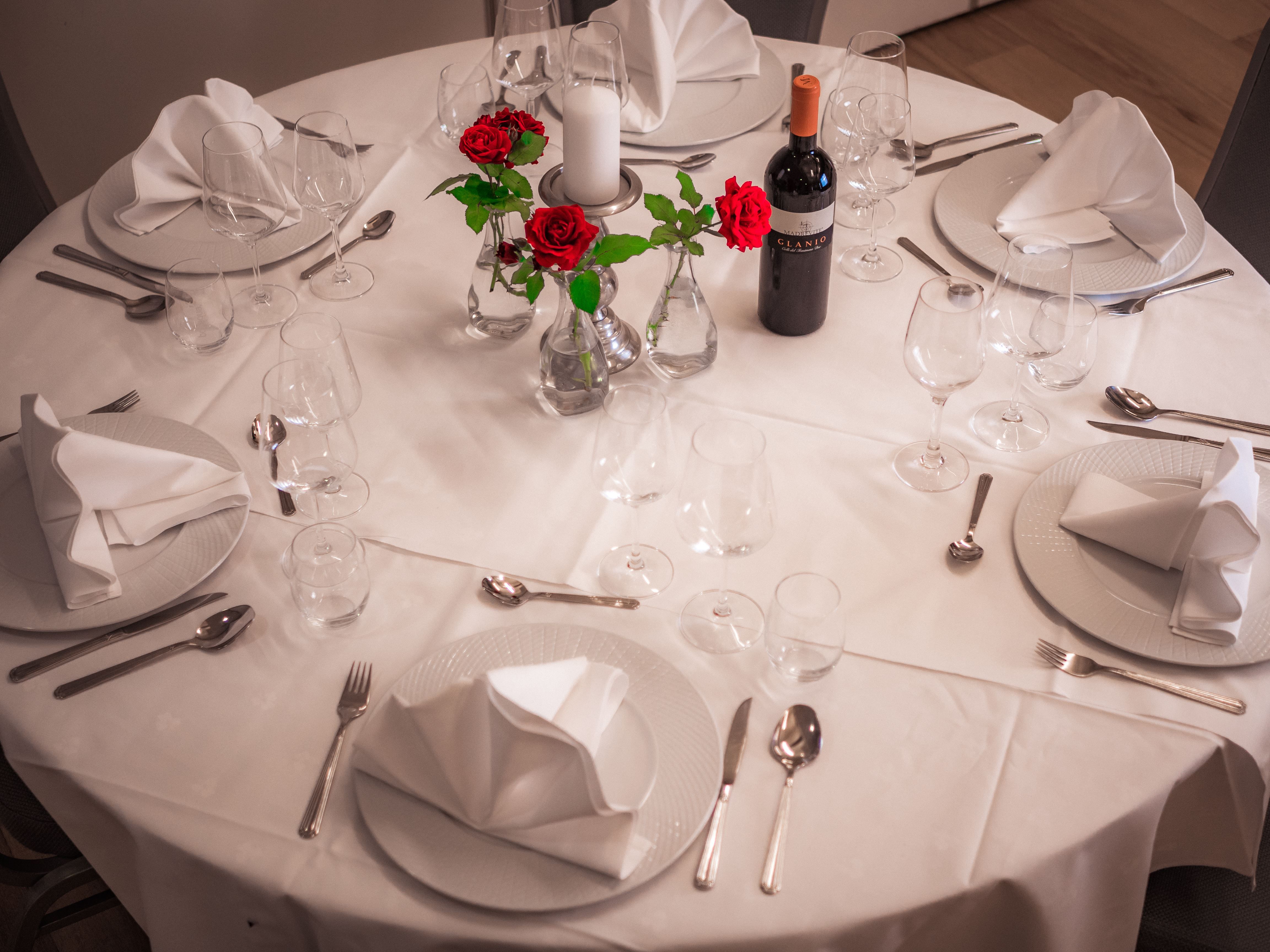 Round table with white tablecloth, folded napkins, wine glasses, cutlery, red roses in vases, candle, and wine bottle.