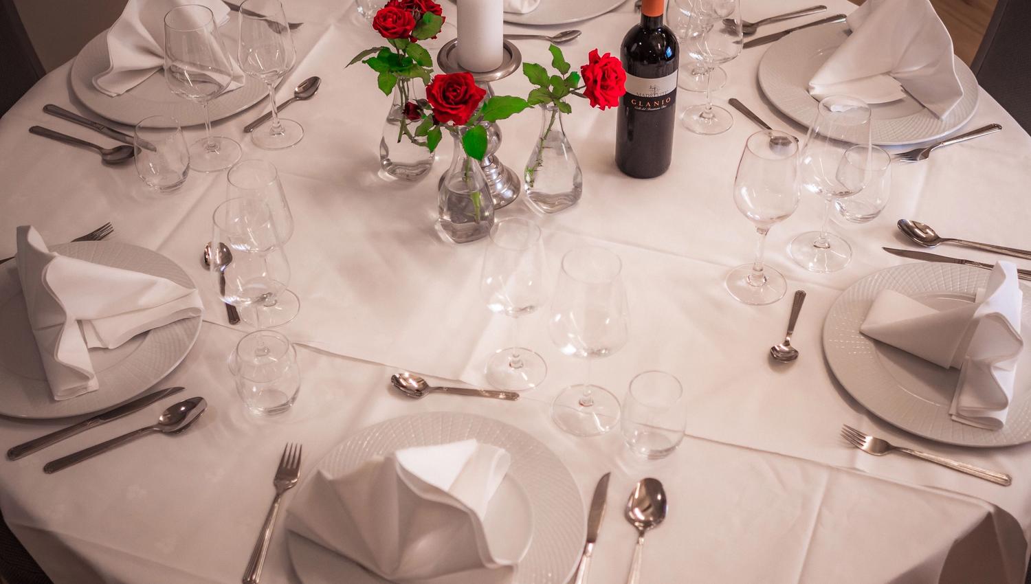 Round table with white tablecloth, folded napkins, wine glasses, cutlery, red roses in vases, candle, and wine bottle.