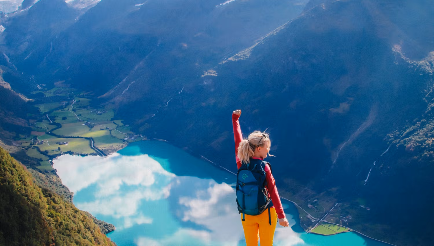 A hiker with a backpack stands on a rocky cliff above a turquoise lake, with mountains and clouds in the distance..