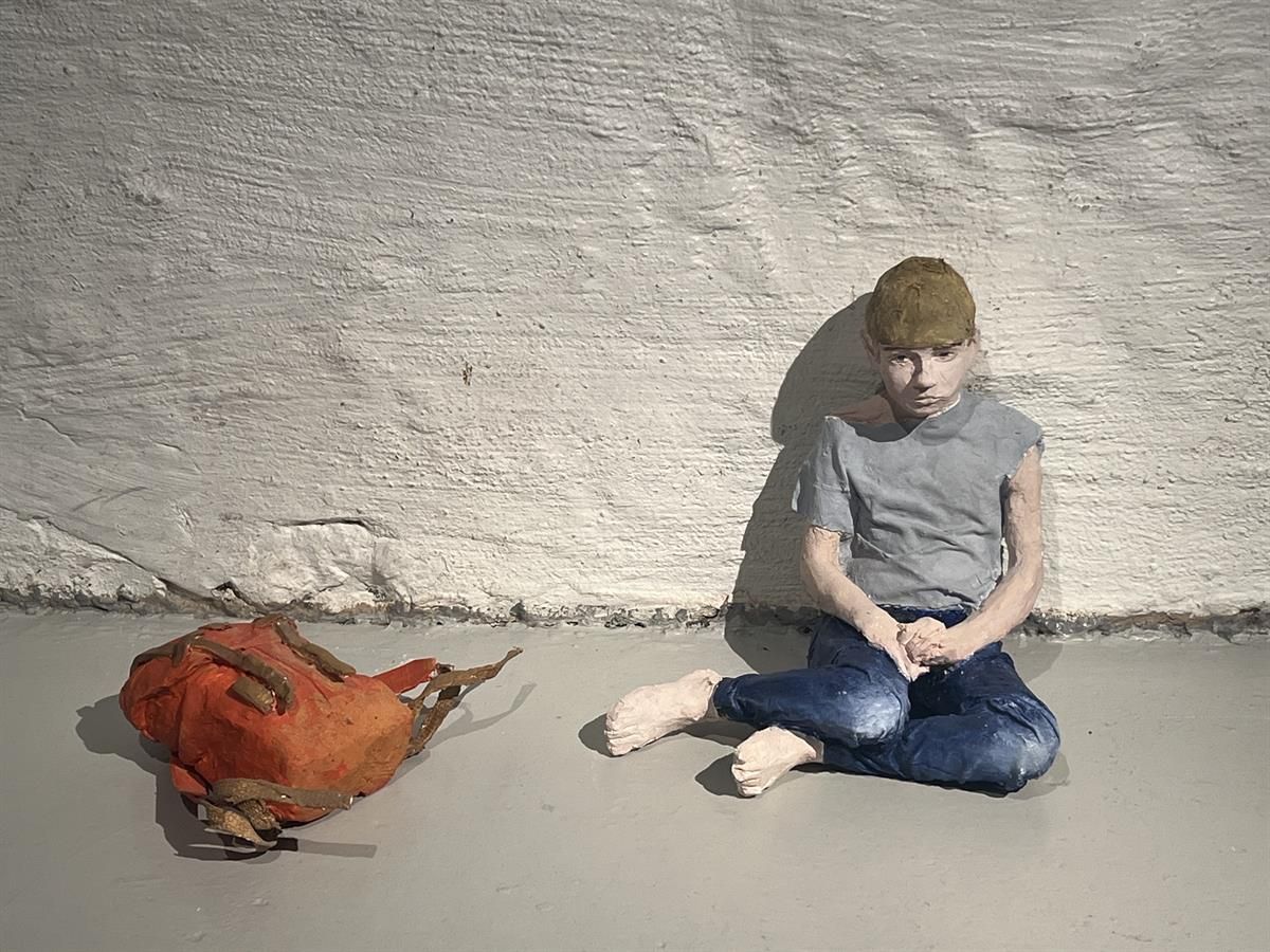 Figure sitting on the floor by a white brick wall with a jacket placed beside.