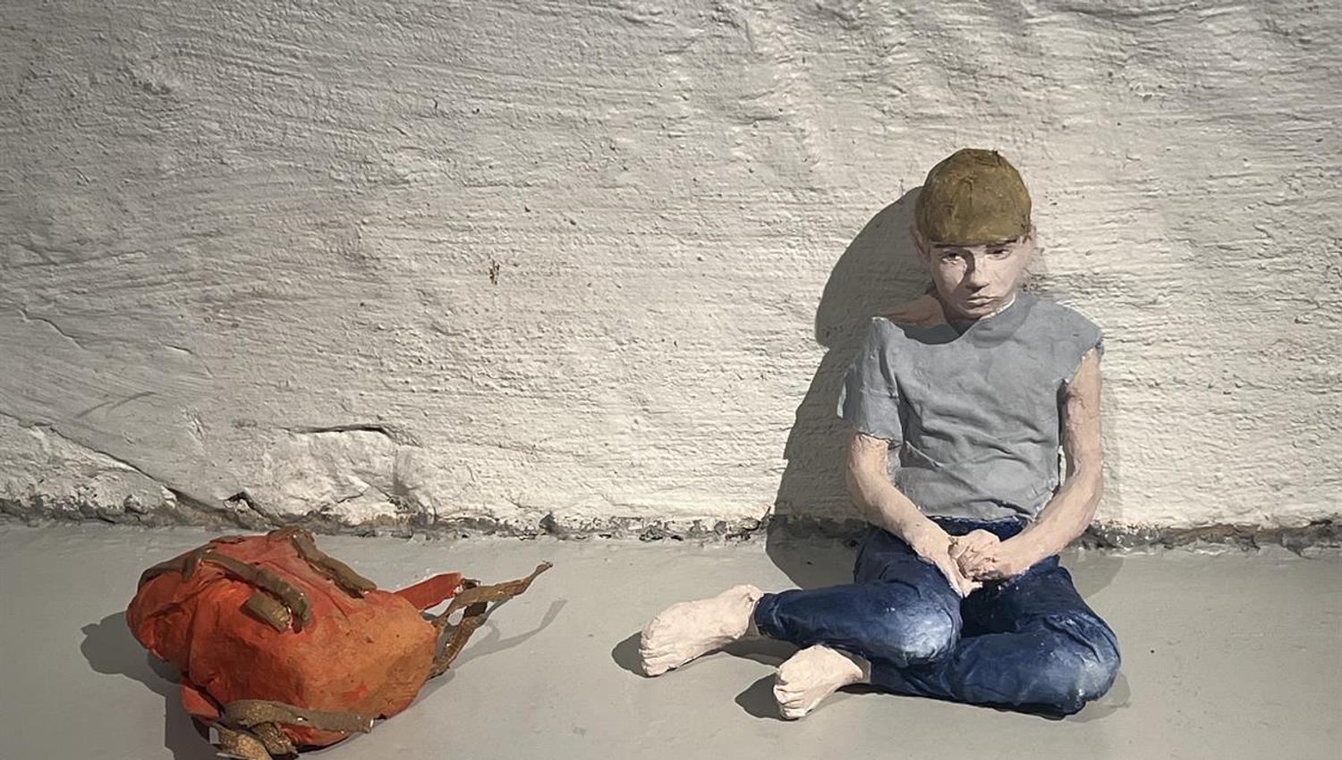 Figure sitting on the floor by a white brick wall with a jacket placed beside.